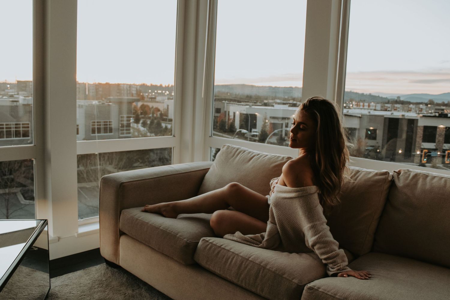 Woman in sweater looking out large windows at city view from modern apartment.
