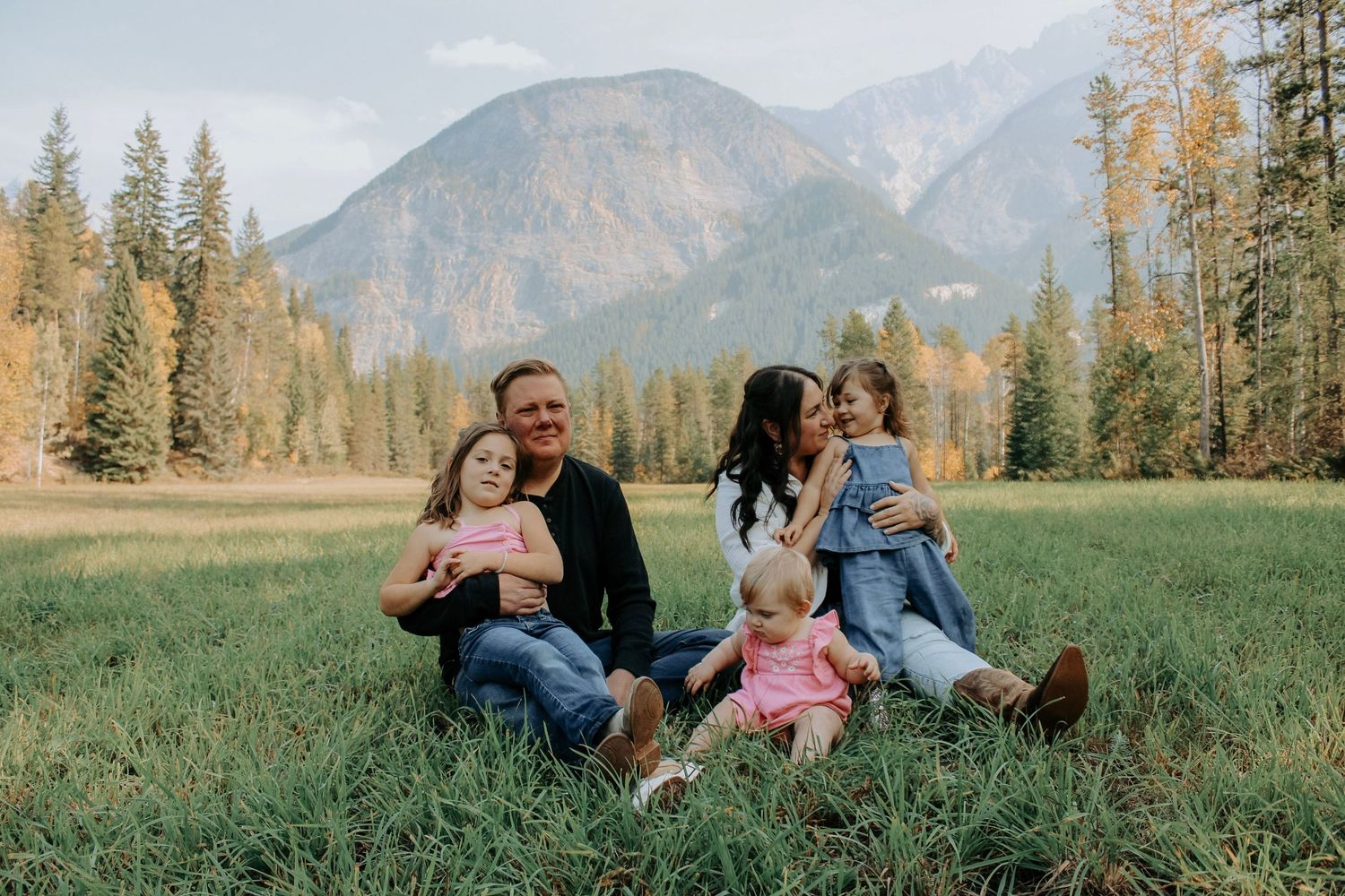 A tender moment shared between two people in a grassy field with mountains and evergreen trees in the background.