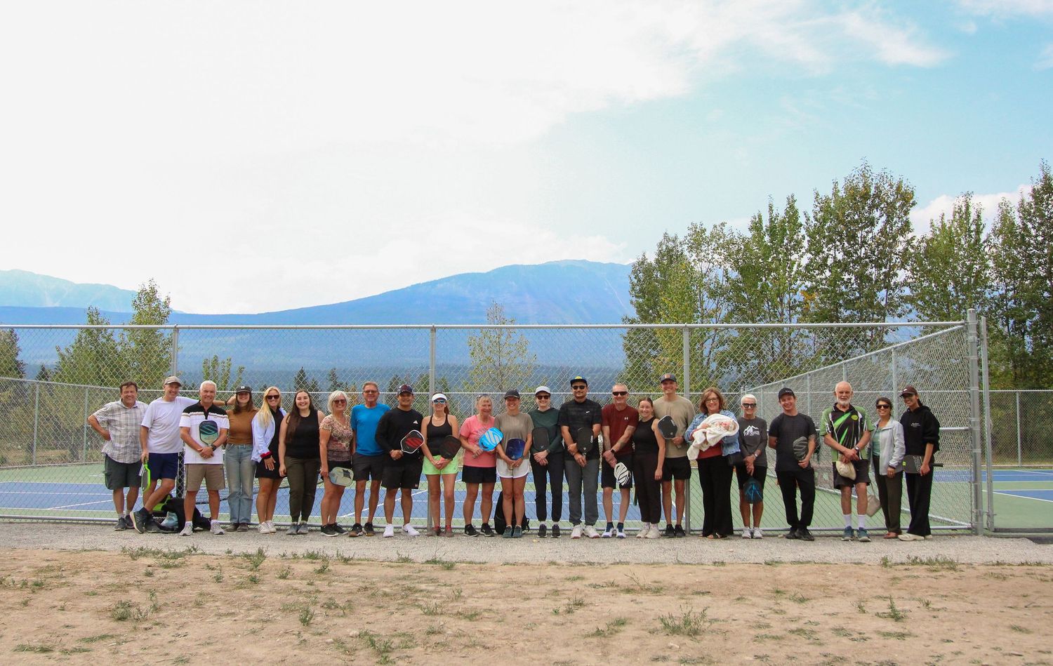 Large group of people posing together on a sunny day near tennis courts with mountain views in the background.