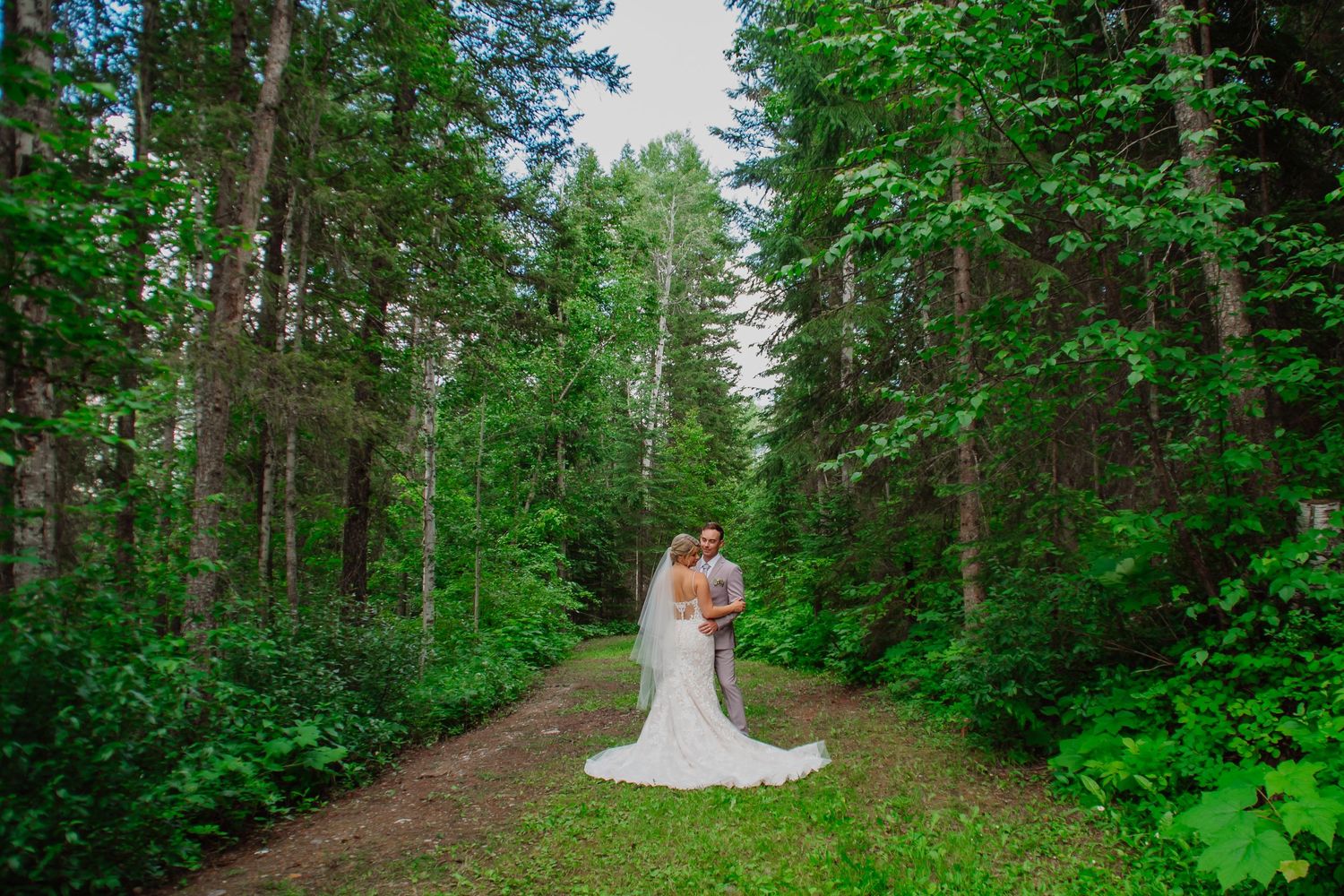 Bride and groom embracing on a forest path surrounded by lush green trees.