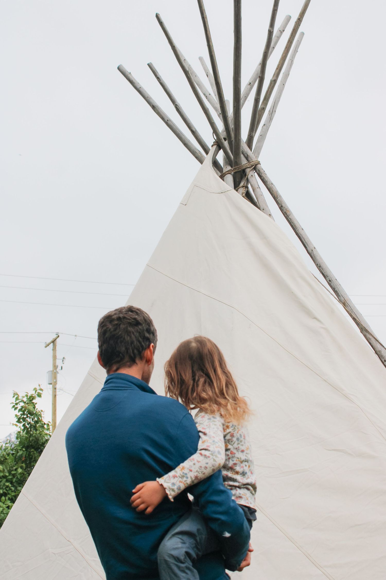 Father holding young child in front of a white canvas teepee with wooden poles against an overcast sky.