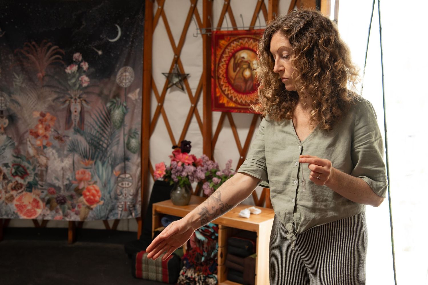 Woman with curly hair holding a pendulum in a bohemian yurt with colorful tapestries and floral decor.