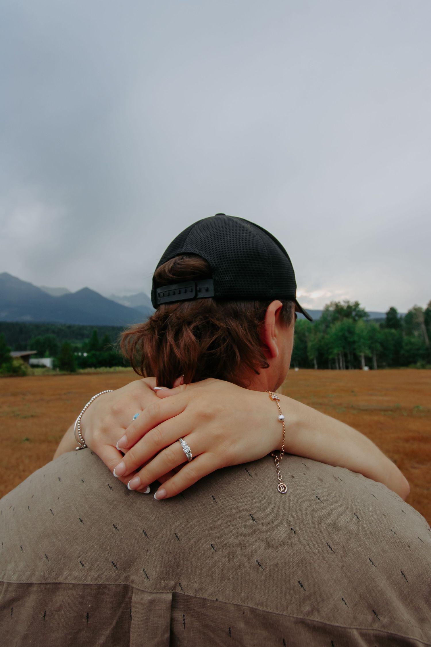 A couple embraces outdoors, woman's hand with ring rests on man's shoulder, mountains visible in background.