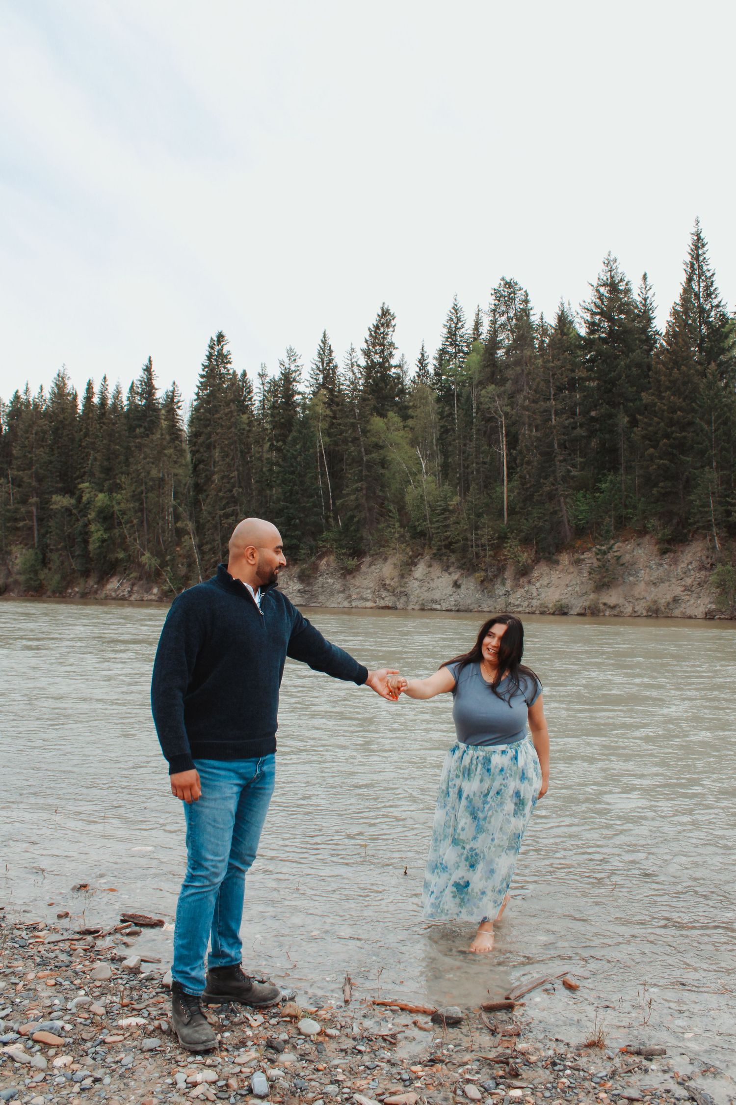 A couple holds hands by a rocky riverbank surrounded by tall evergreen trees, as the woman wades in shallow water.