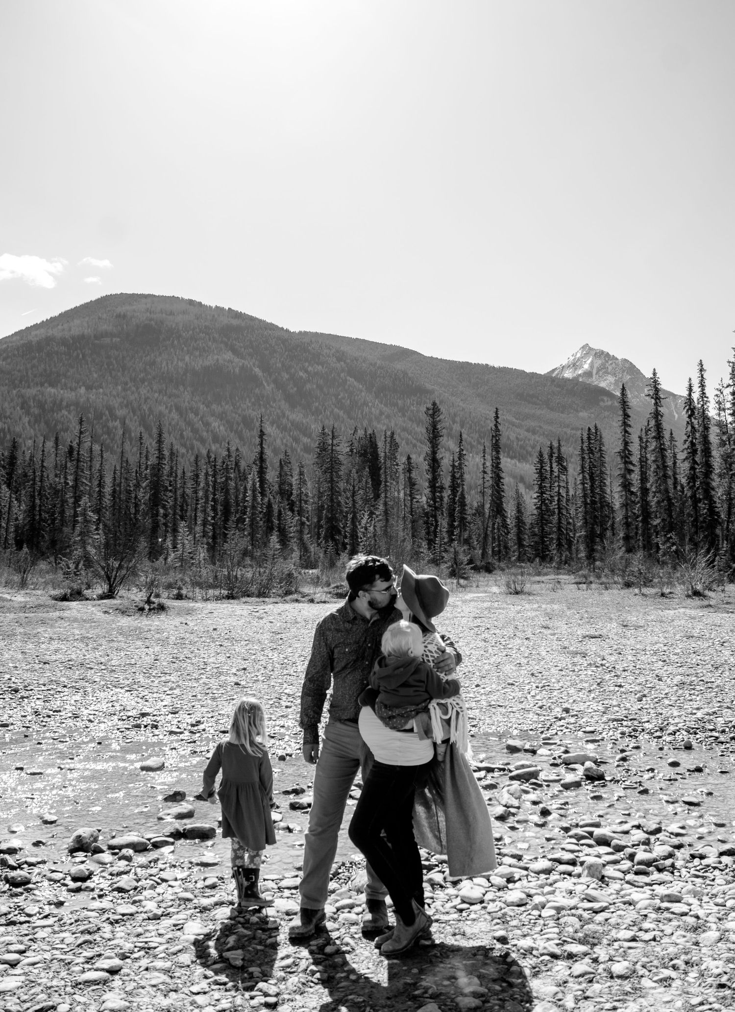 Black and white photo of a family embracing on a rocky riverbed with mountains and pine trees in the background.