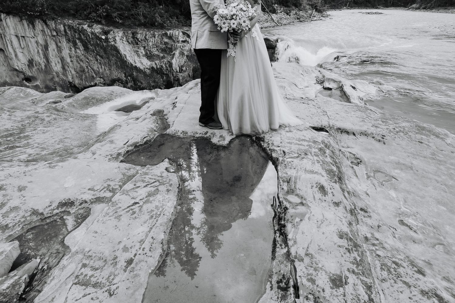 Black and white wedding photo of bride and groom standing on rocky shore holding bouquet.