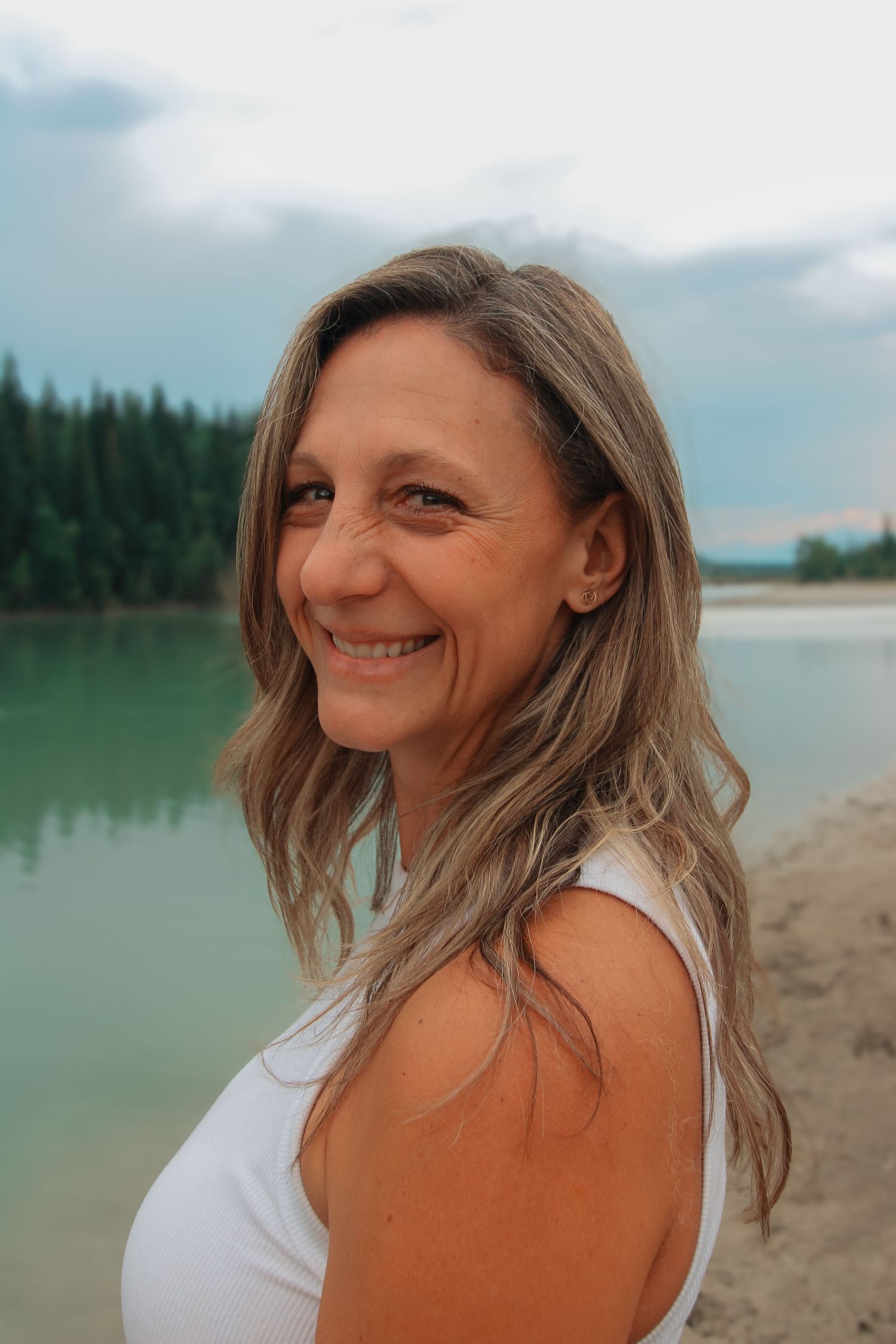 Smiling middle-aged woman with long blonde hair poses by a serene lake with evergreen trees in the background.