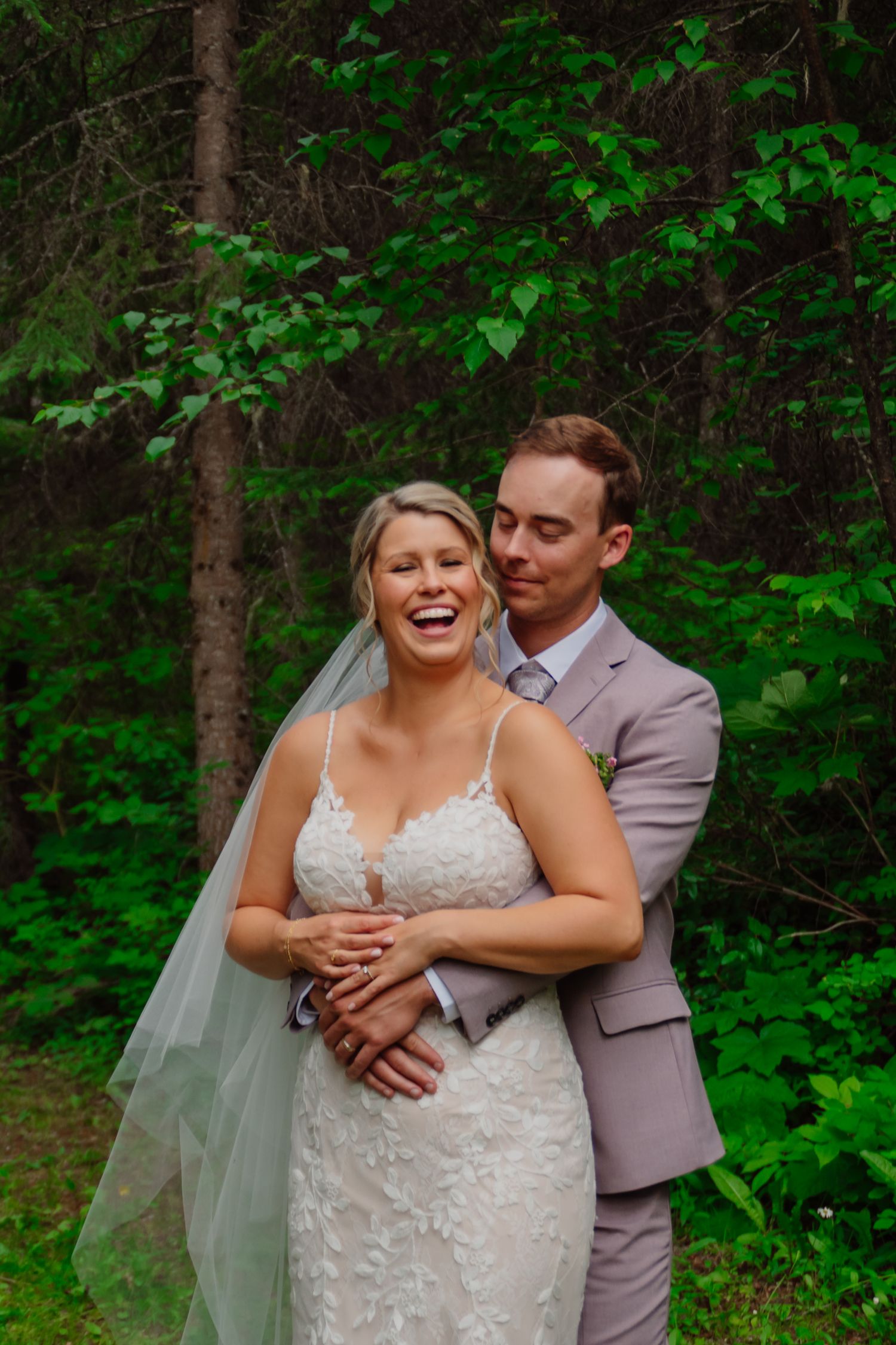 Bride in lace gown laughing joyfully as groom in blush suit embraces her from behind in a lush green forest.