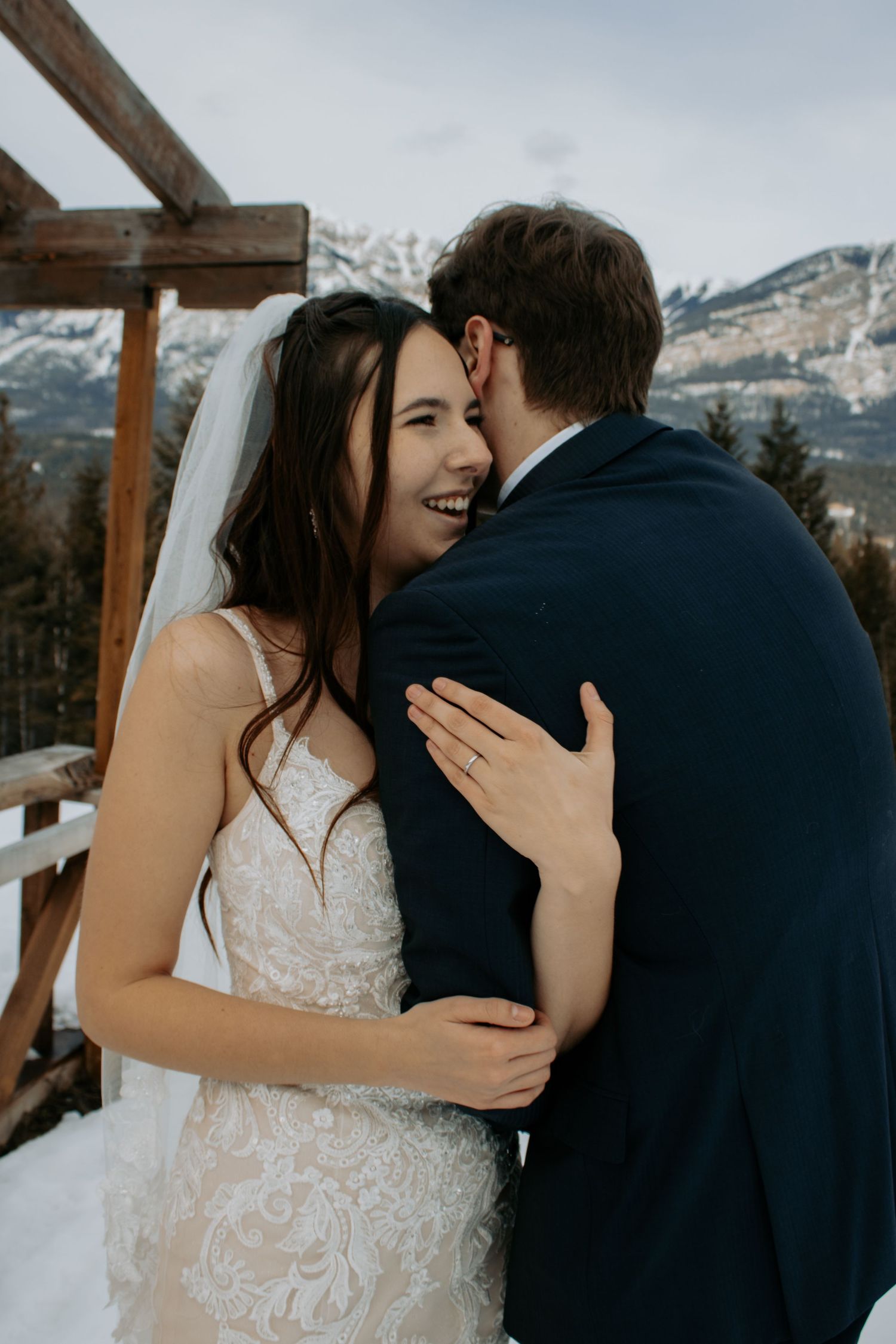 Bride in lace gown and veil embraces groom in navy suit on snowy mountain backdrop.