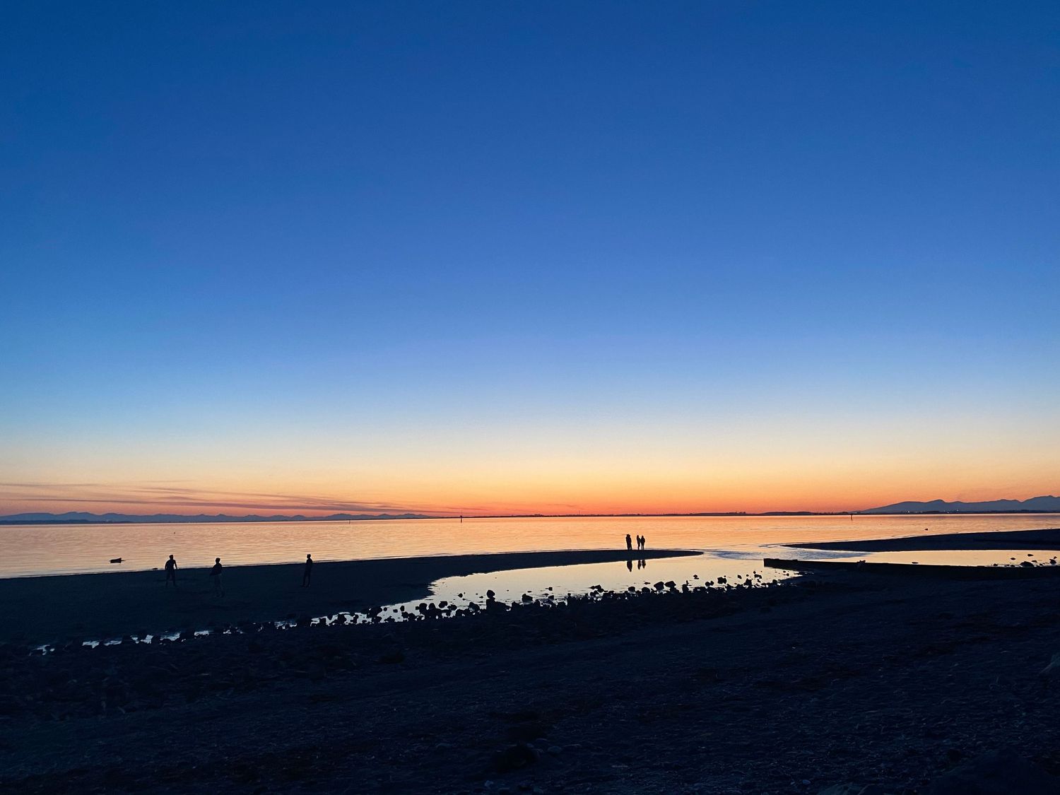 Silhouettes of people walking along a beach at sunset with vibrant orange and blue sky reflecting on calm waters.