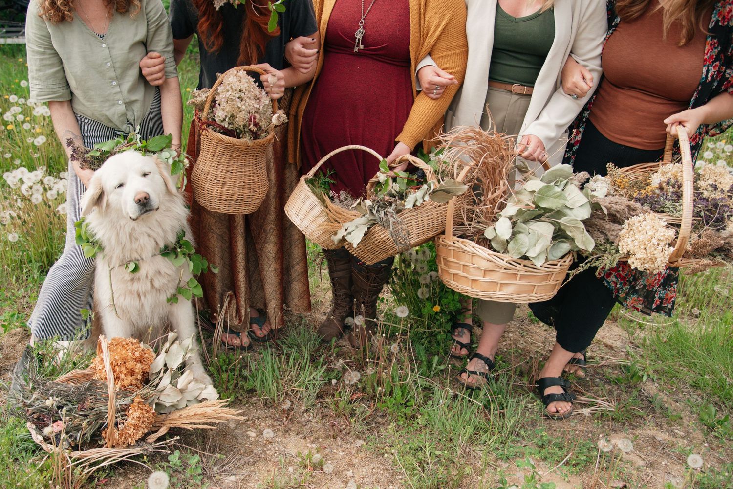 Women holding wicker baskets filled with dried flowers and foliage outdoors, with a fluffy white dog sitting beside them.