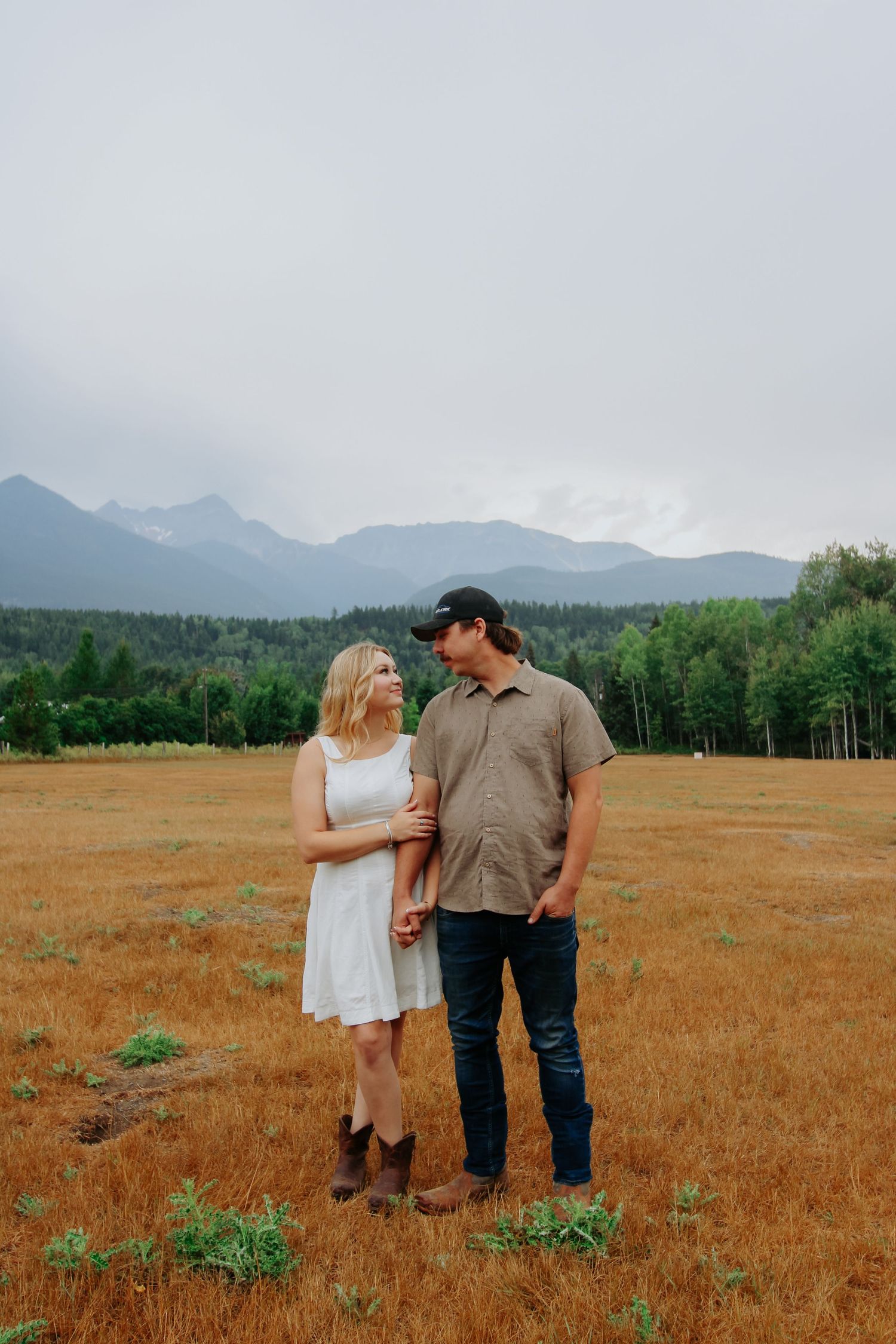 Couple holding hands in golden field with mountain backdrop, woman in white dress and cowboy boots, man in cap and jeans.