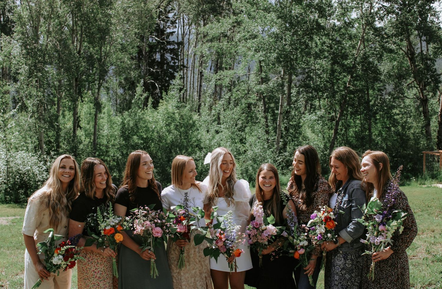 A group of bridesmaids in floral dresses holding bouquets of flowers in a lush, green forest setting.
