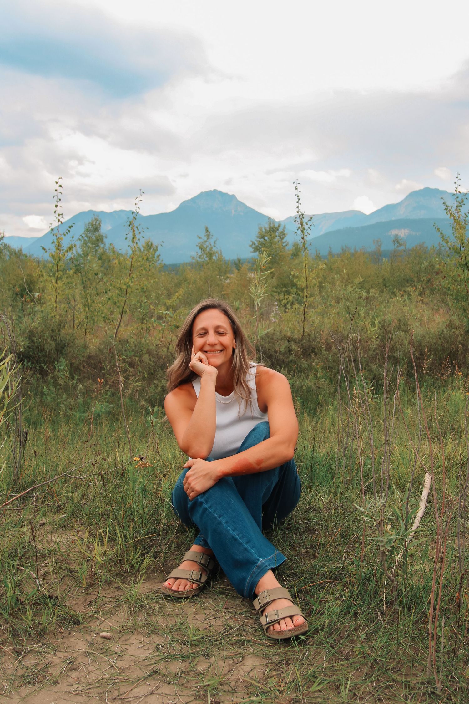 A smiling woman sits cross-legged on a dirt path in a green meadow with mountains and cloudy sky behind her.