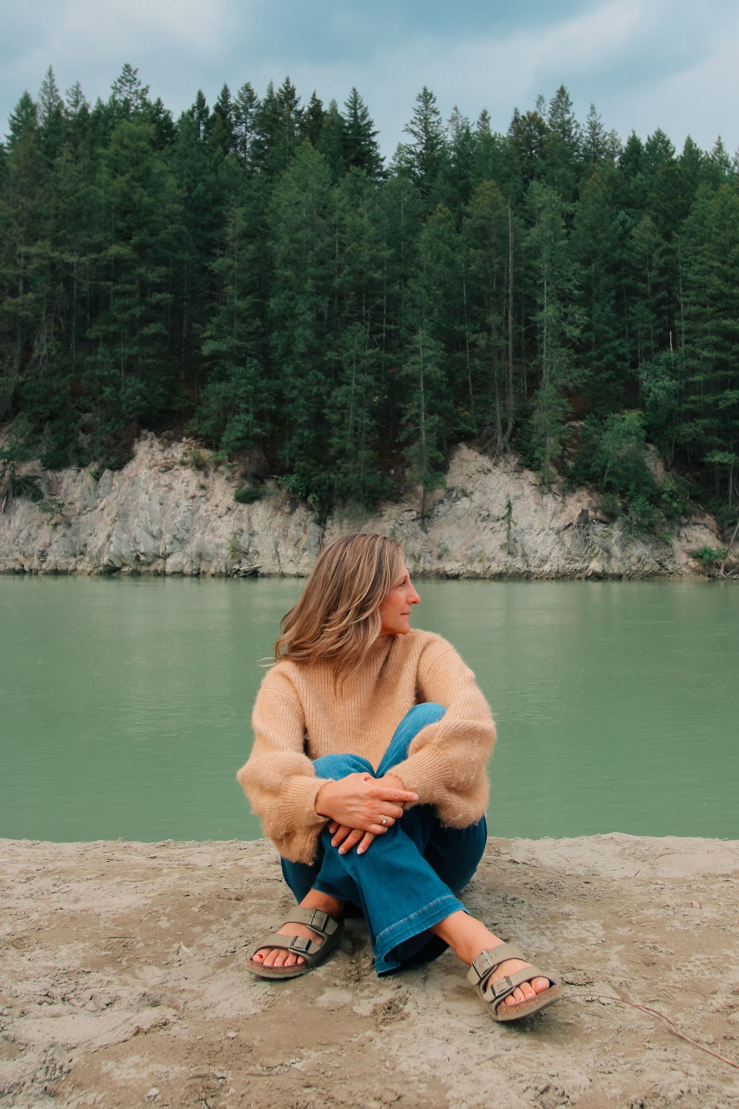 Woman in beige sweater and blue jeans sits on rocky shore beside turquoise lake with pine forest backdrop.