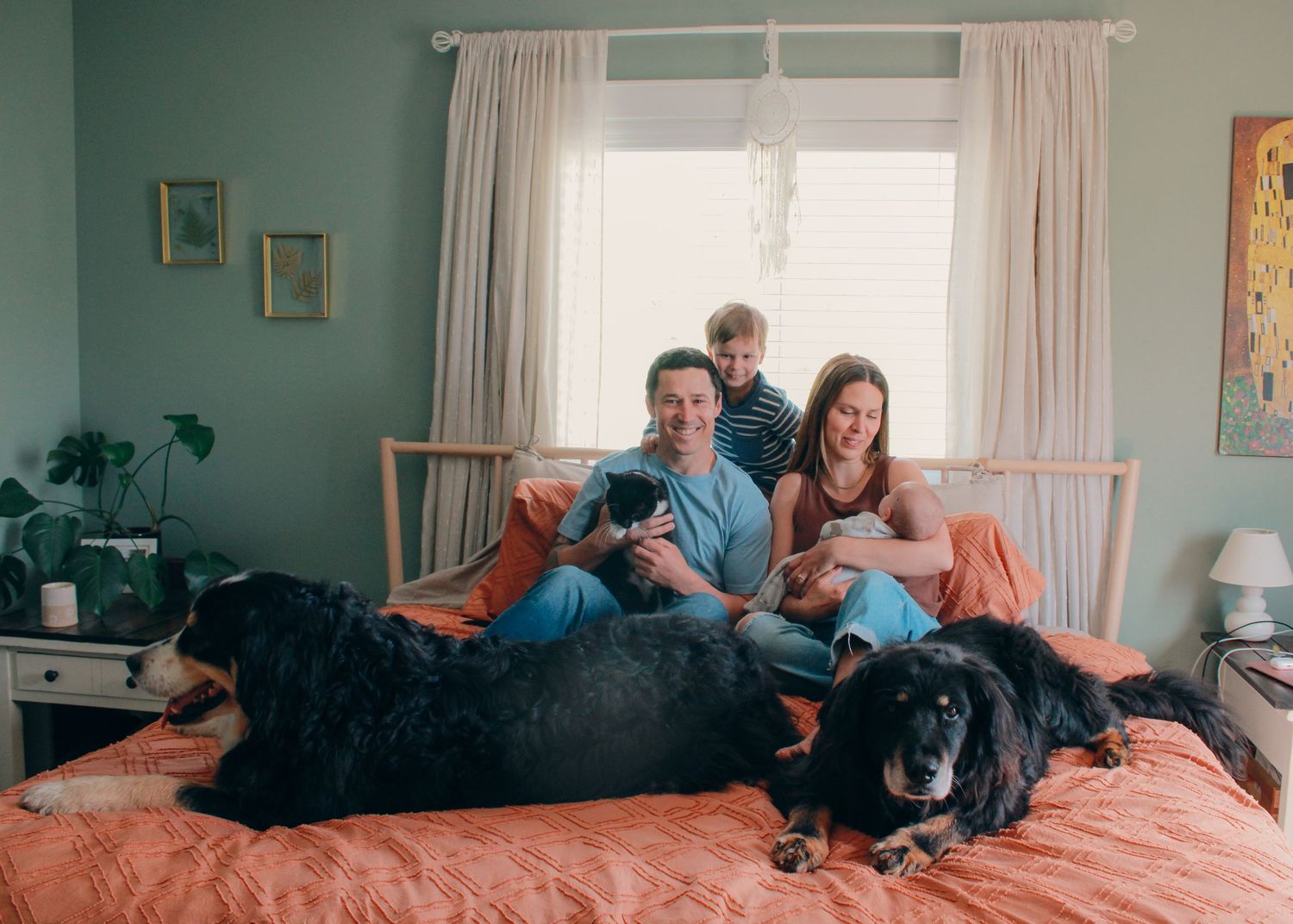 Family of three with two black dogs relaxing on an orange bed in a cozy teal bedroom near a bright window.