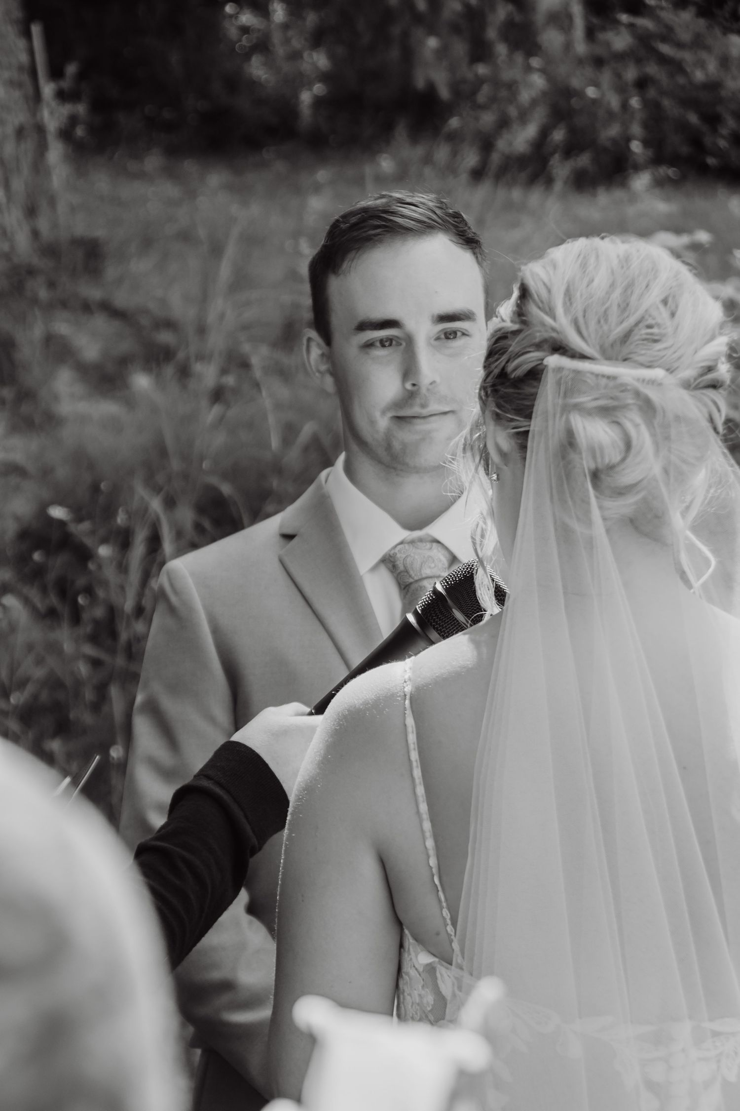 Black and white wedding photo of groom in suit facing bride in veil during outdoor ceremony.