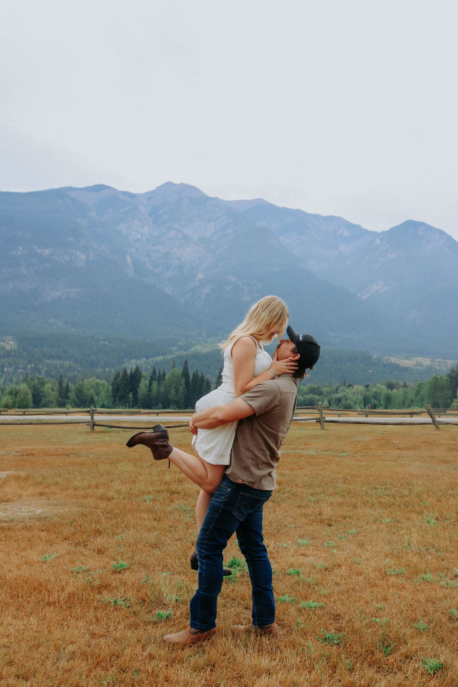 A man lifts a woman in a romantic embrace in a golden field with mountains in the background.