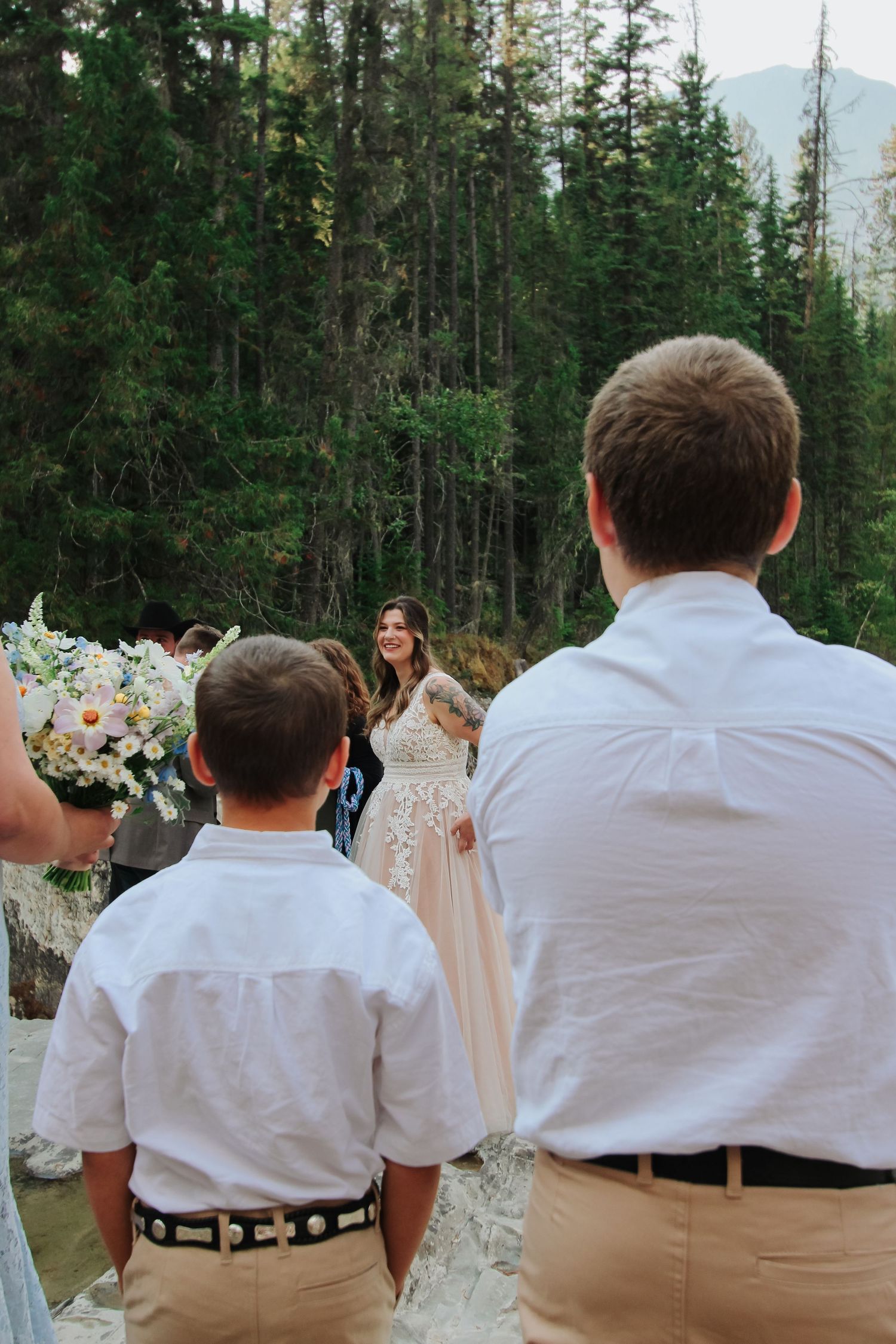Bride in lace gown smiles at guests during outdoor forest wedding ceremony with floral arrangements.