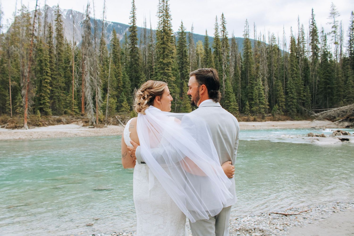 Wedding couple walks along a scenic mountain lake surrounded by evergreen trees.