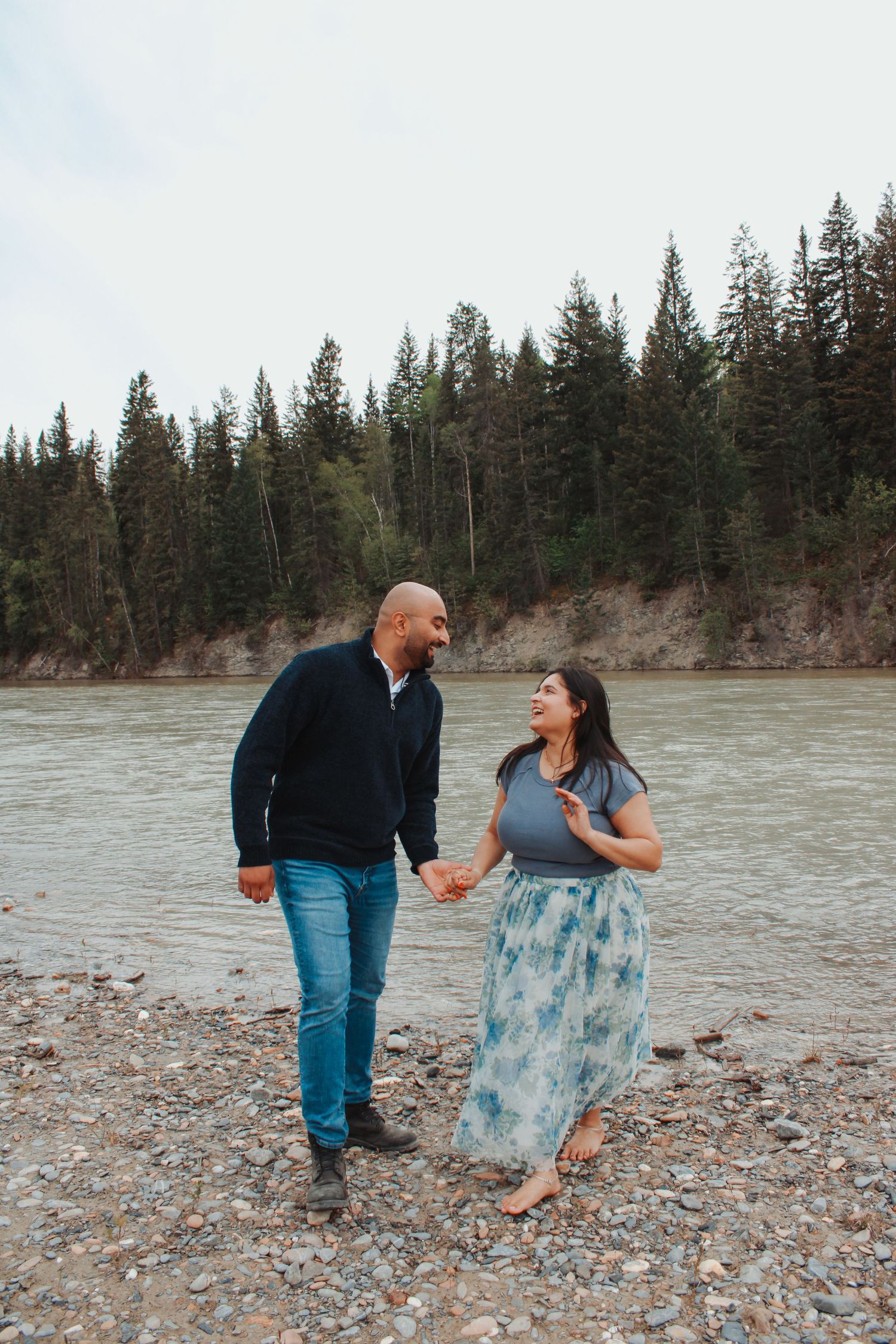 Couple holding hands and smiling at each other on a rocky riverbank surrounded by tall pine trees.