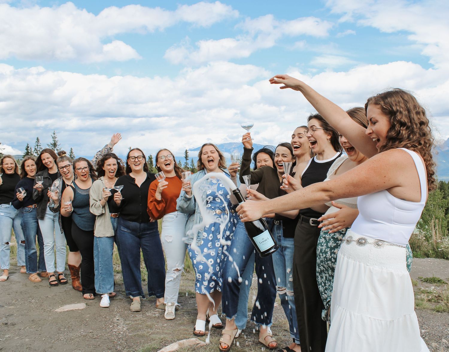Group of women celebrating outdoors, popping champagne bottle, laughing joyfully under blue sky with mountains behind.