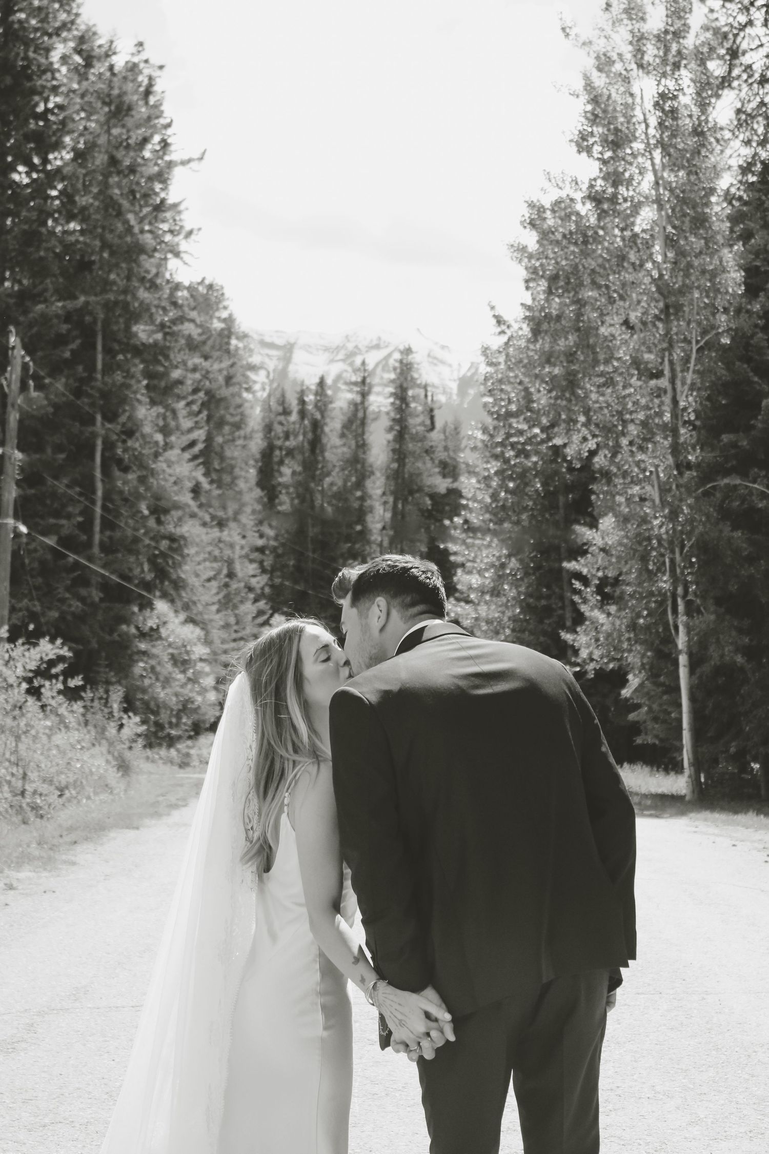 Black and white photo of a bride and groom holding hands, walking down a forest road with mountain views.