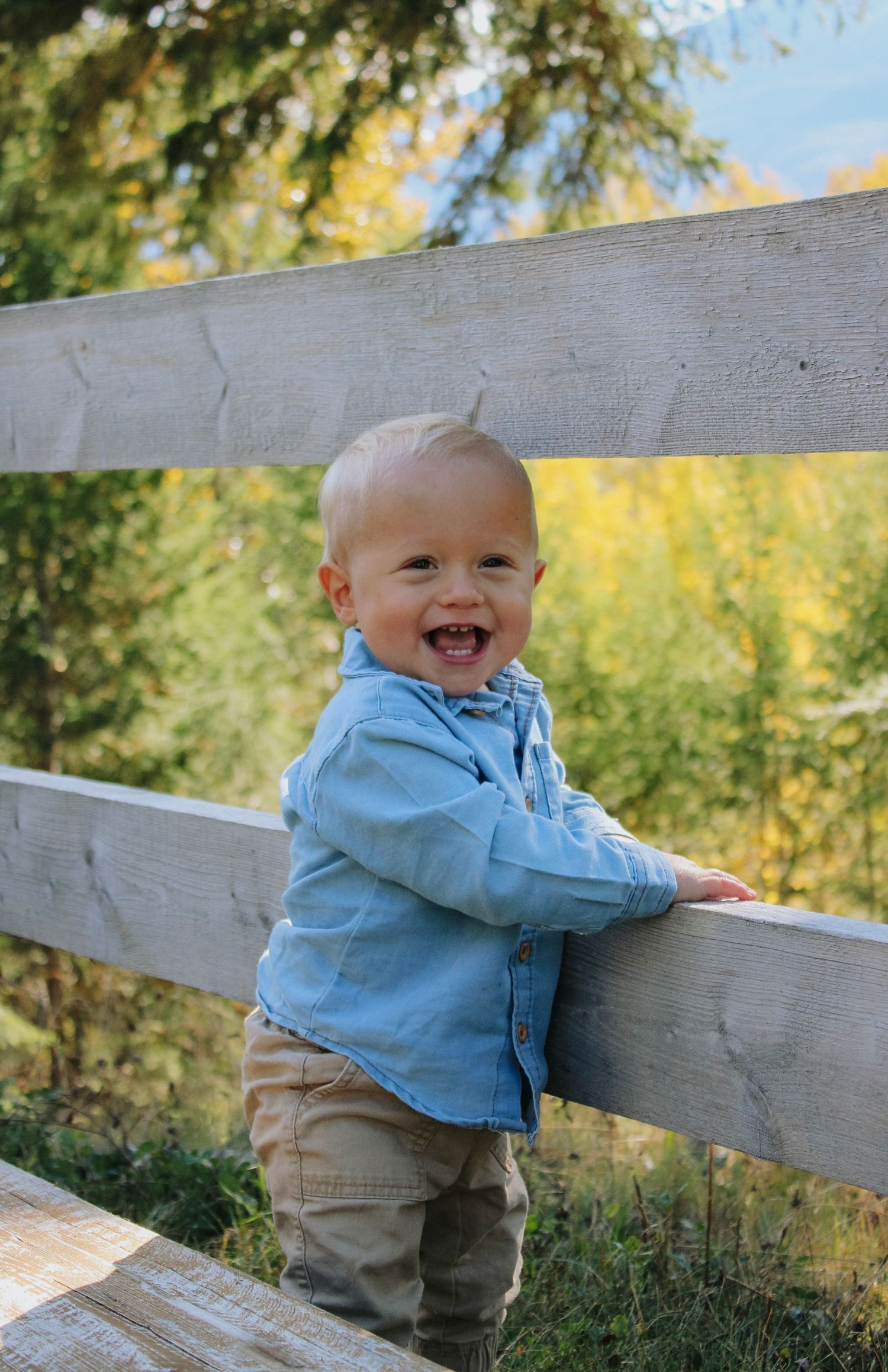 A toddler in a blue jacket and khaki pants stands playfully by a wooden fence in an outdoor setting.