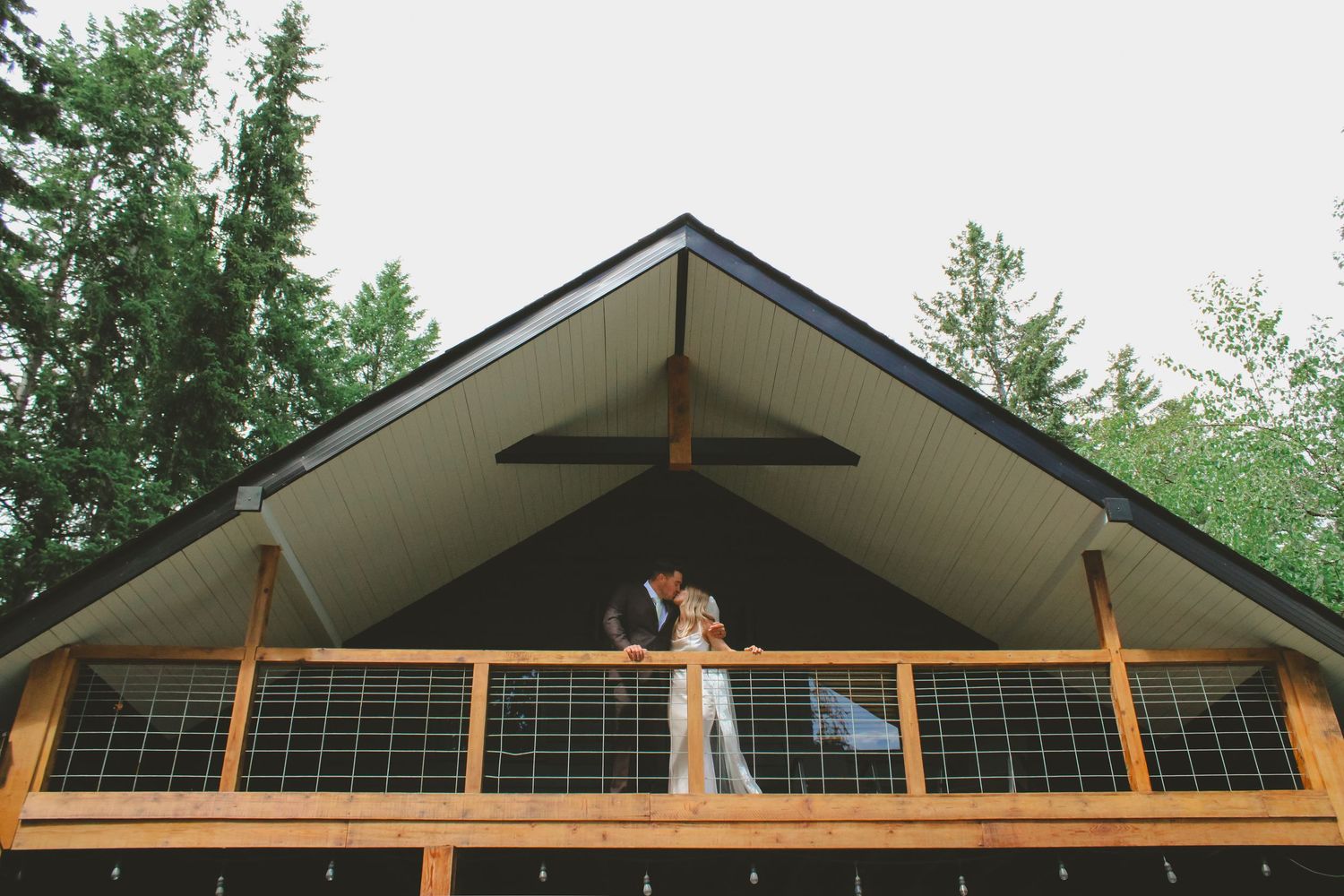 Bride and groom share a romantic moment on a wooden chalet balcony surrounded by tall green trees.