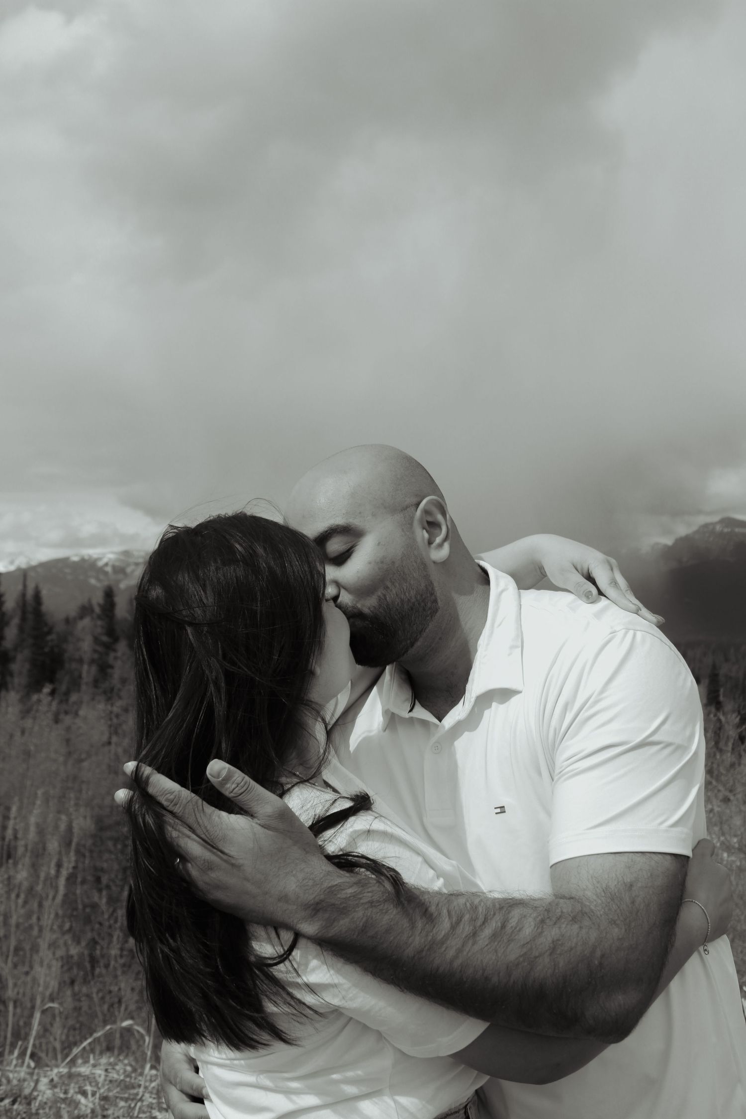 Black and white photo of a bald man kissing a woman with long dark hair outdoors with mountains in the background.