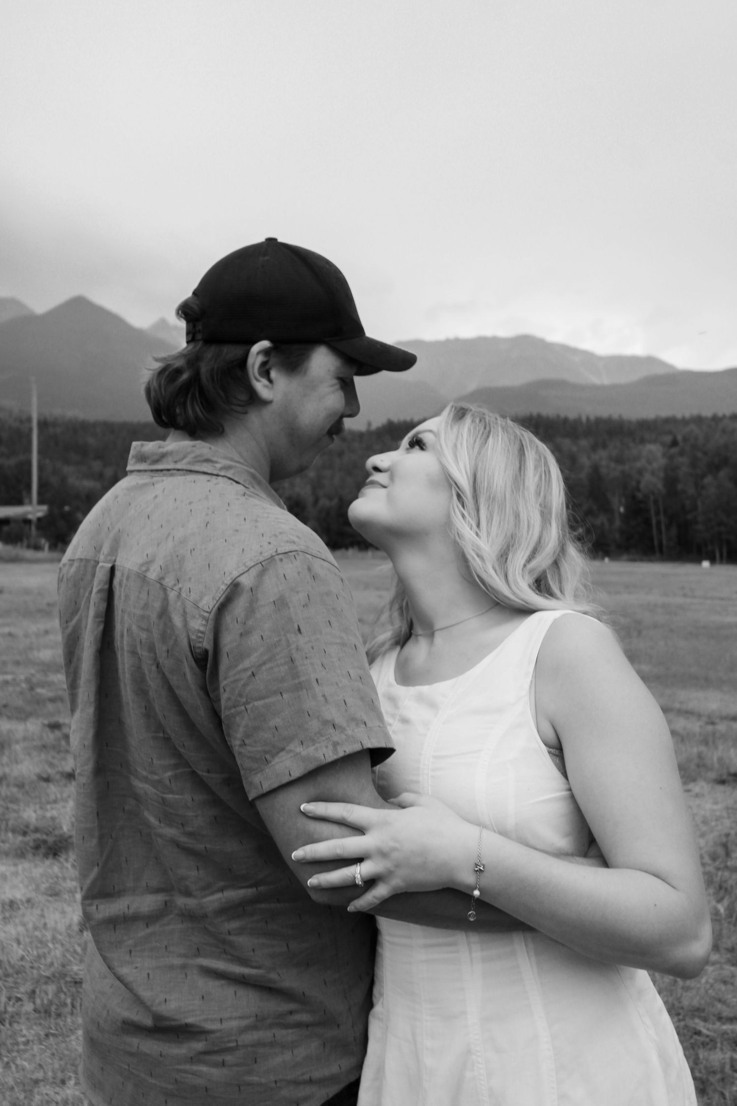 Black and white photo of couple gazing at each other in field with mountain range visible in background.