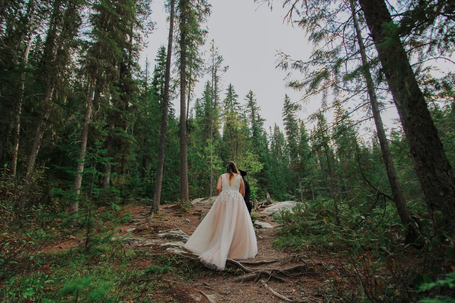 Bride in white gown walking through lush green forest on a woodland trail during an outdoor wedding.