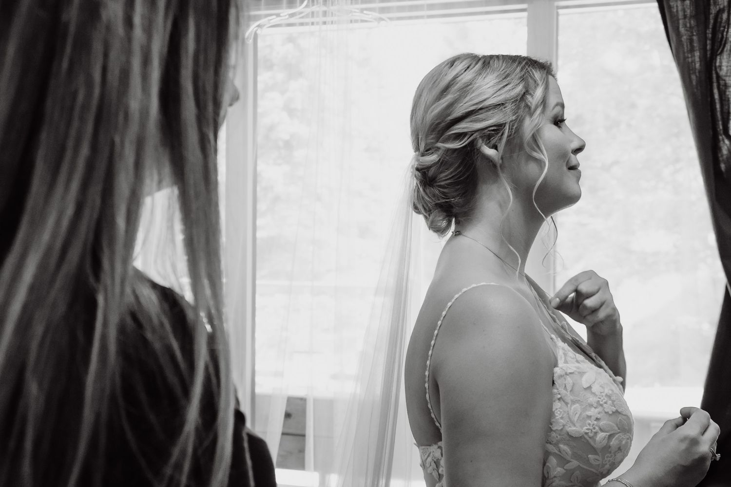 Black and white photo of a bride in a lace gown and veil gazing out a window while being helped by bridesmaids.