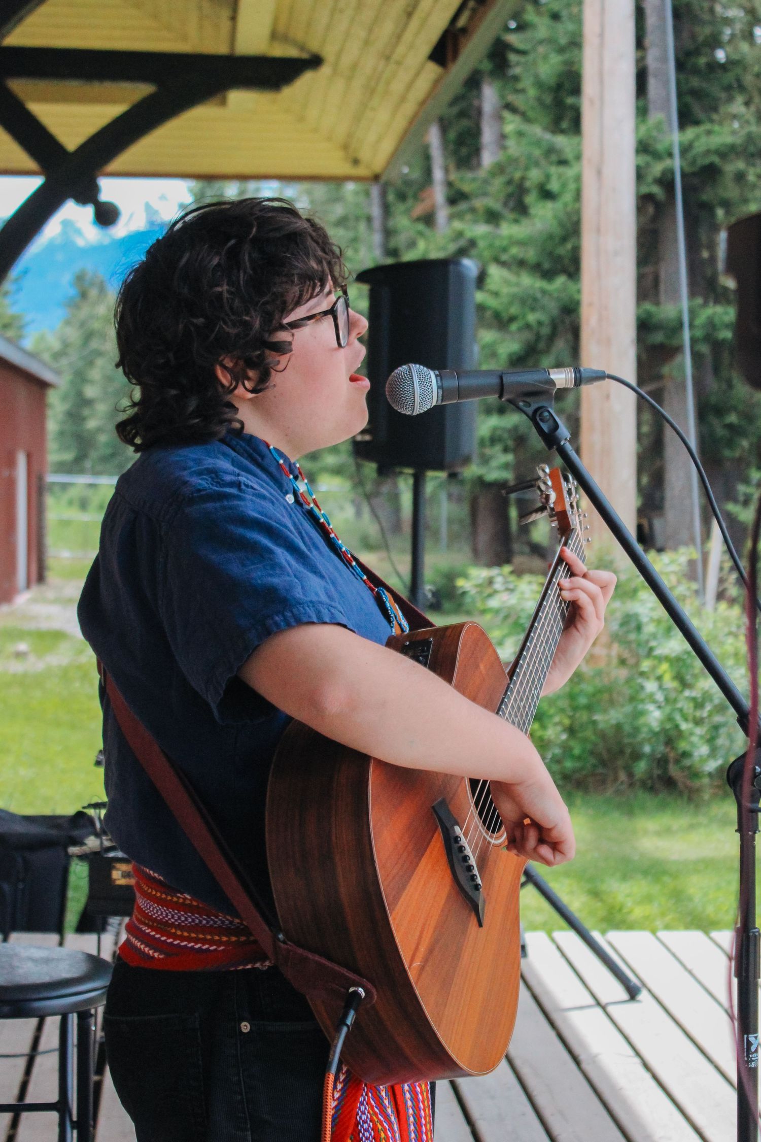 Young musician with glasses playing acoustic guitar and singing into microphone on outdoor stage.