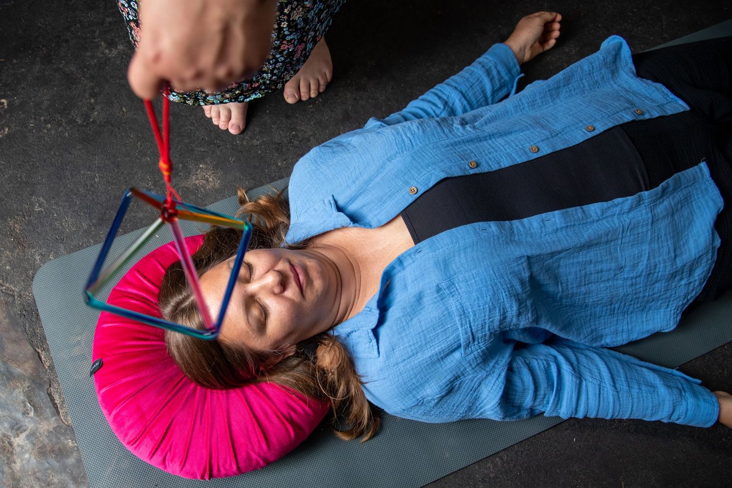 Woman lying on yoga mat with pink neck pillow, wearing blue shirt, with colorful string figure held above her face.