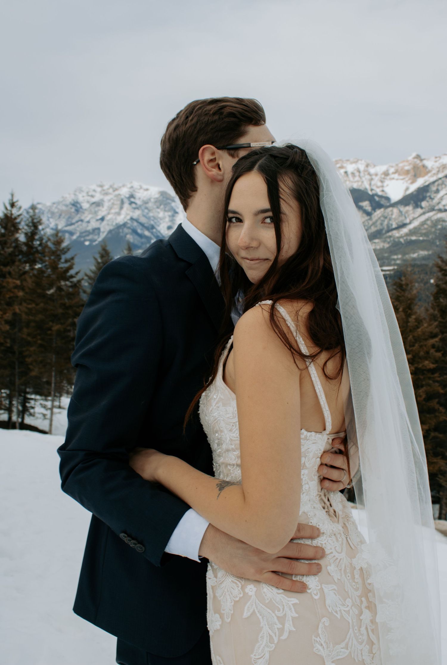 Bride and groom embrace in snowy mountain landscape, bride in lace gown with veil, groom in navy suit.