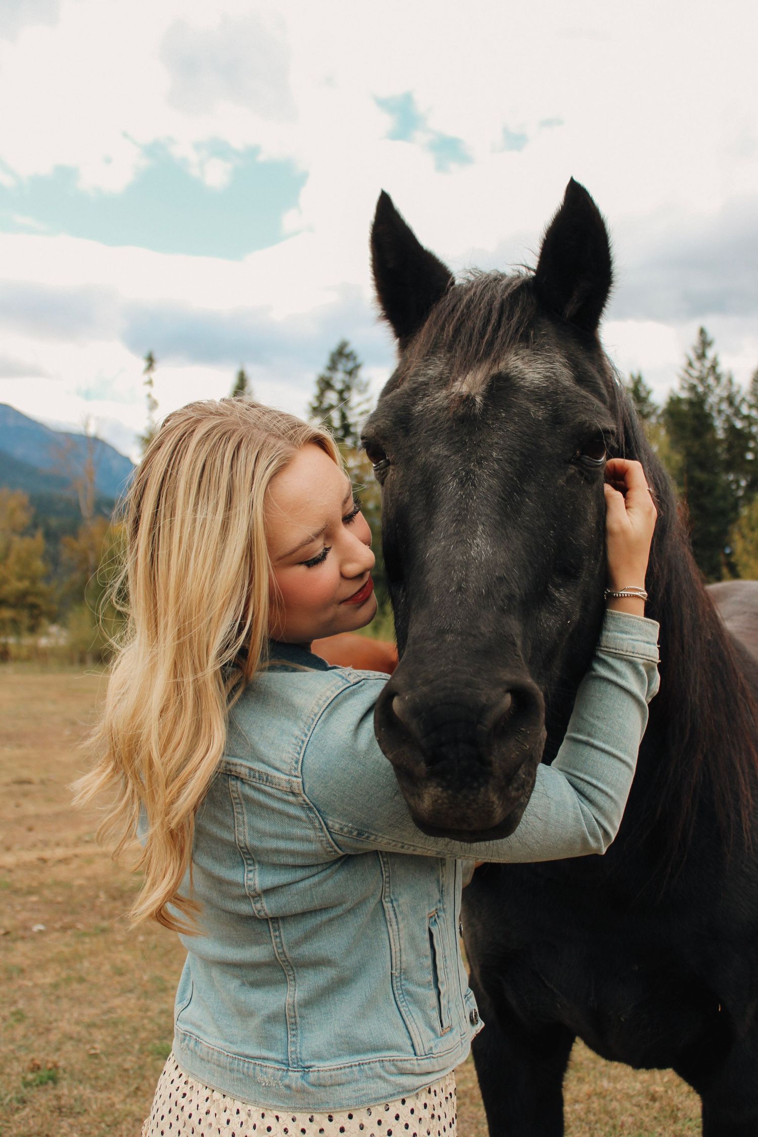Woman in denim jacket sharing an affectionate moment with black horse in mountain field.