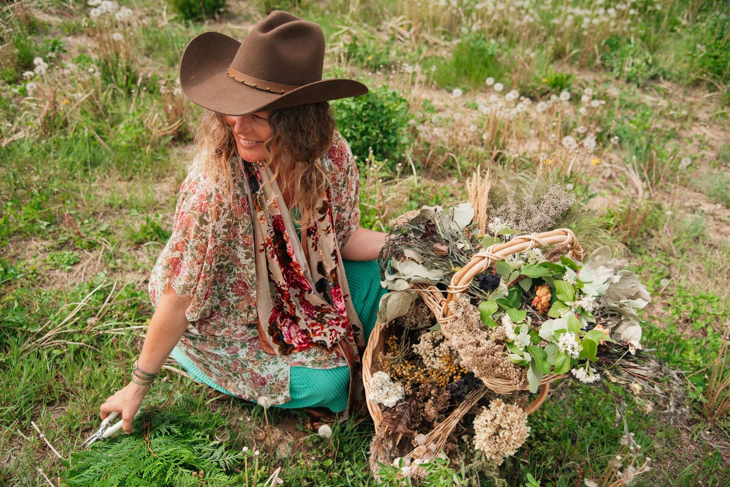 Woman in floral dress and brown cowboy hat sits outdoors arranging a rustic basket of wildflowers and natural elements.