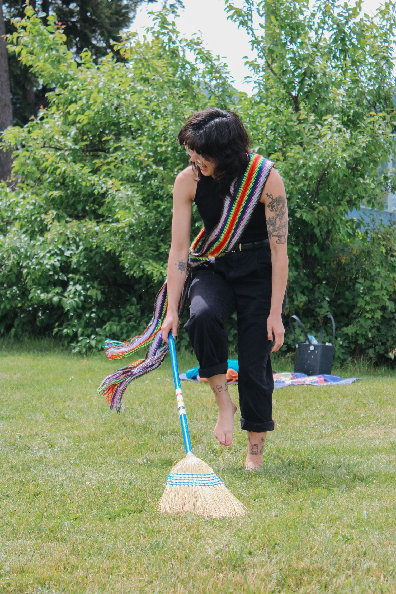 Barefoot woman in black outfit with rainbow sash holds a broom on green lawn surrounded by lush garden.