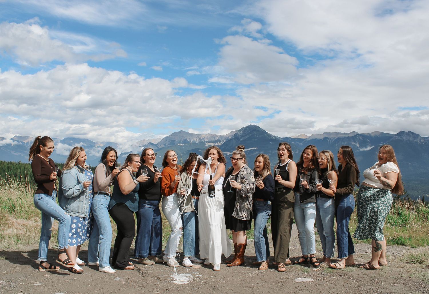 Group of women celebrating outdoors with mountain backdrop under blue skies, laughing and holding drinks.