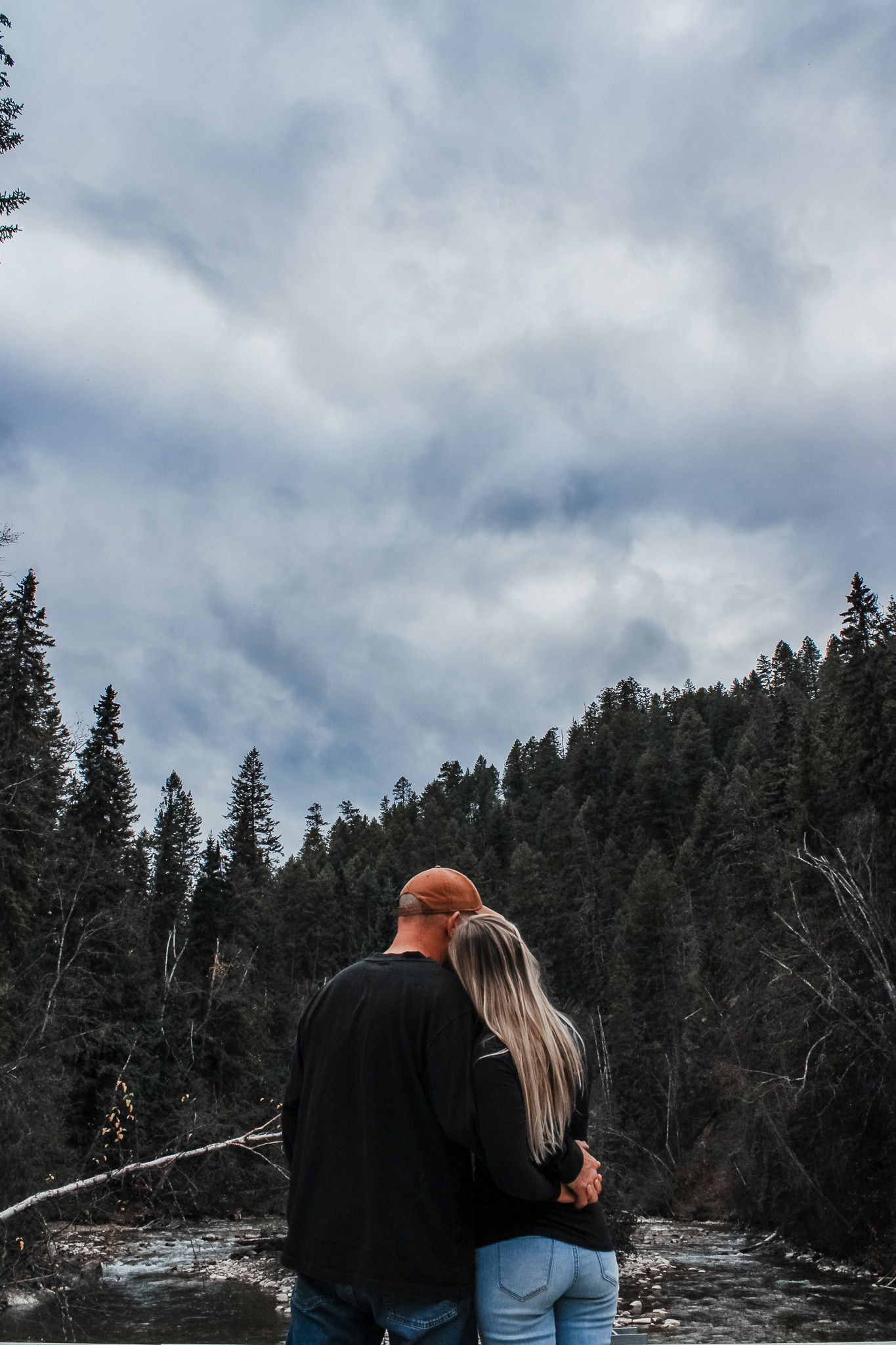 Couple stands on snowy hillside overlooking evergreen forest under cloudy sky.