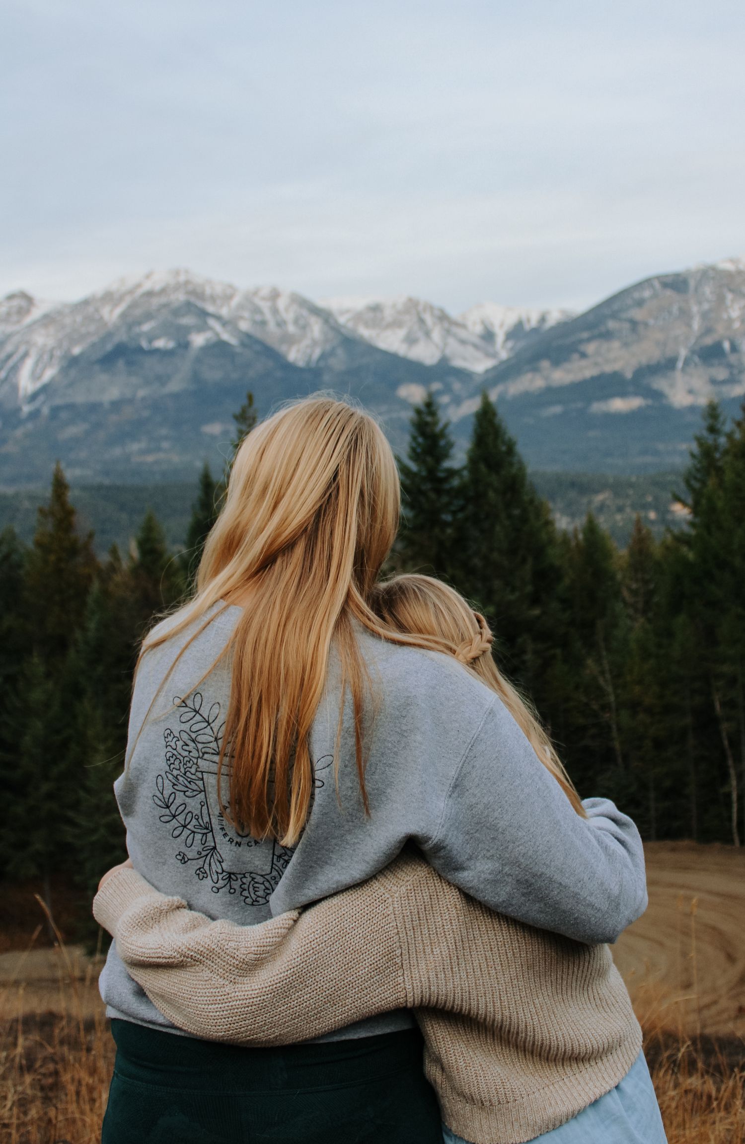 Two people in matching grey sweaters embrace while looking at mountain views.