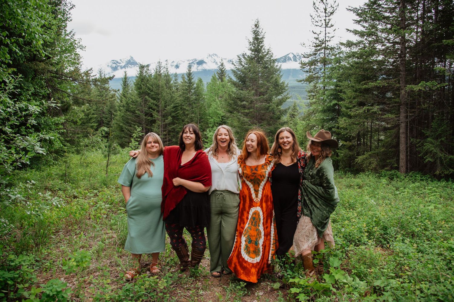 Six women smiling together in a lush forest clearing with snow-capped mountains in the background.