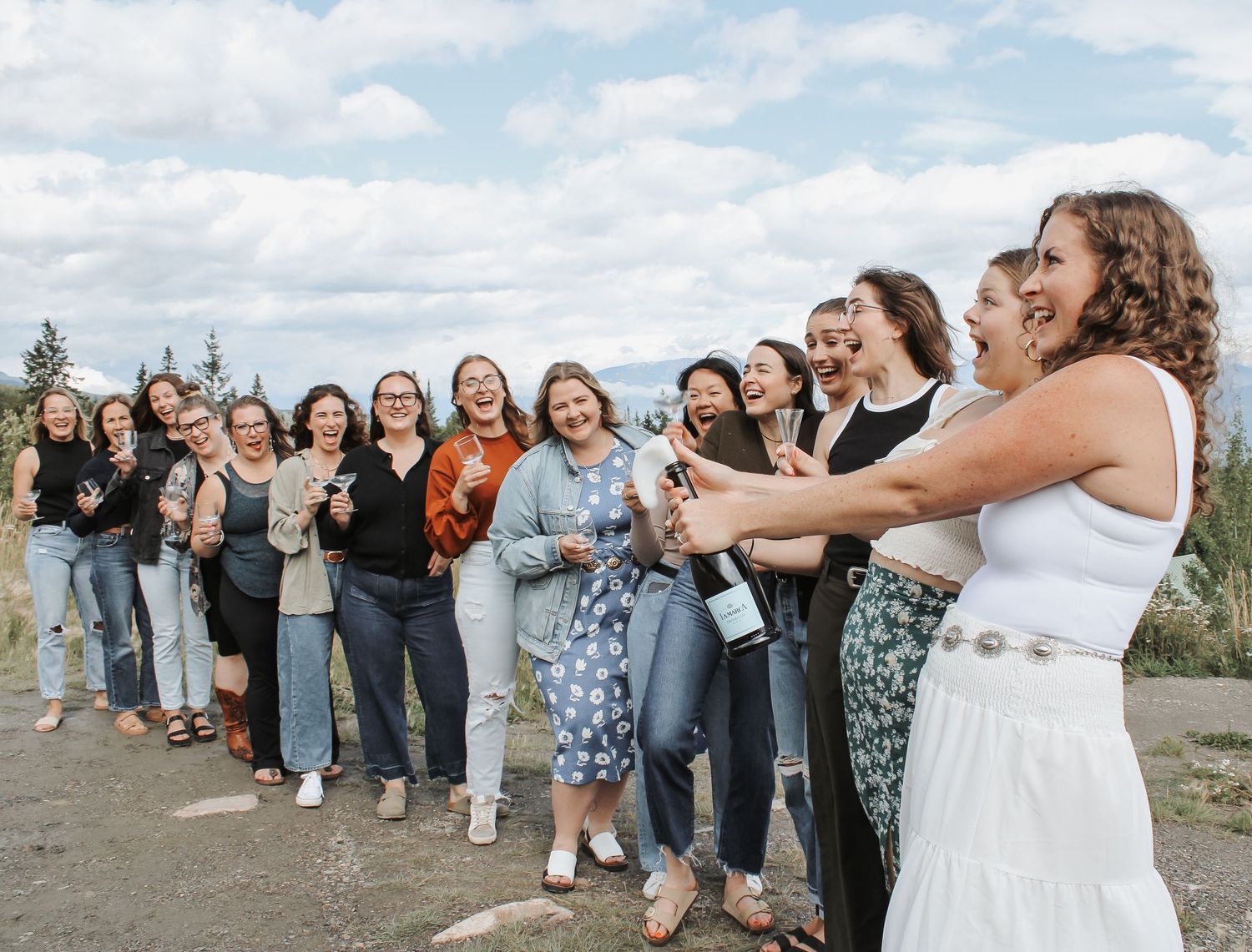 Group of women laughing as bride pops champagne bottle outdoors at scenic overlook during bachelorette party.