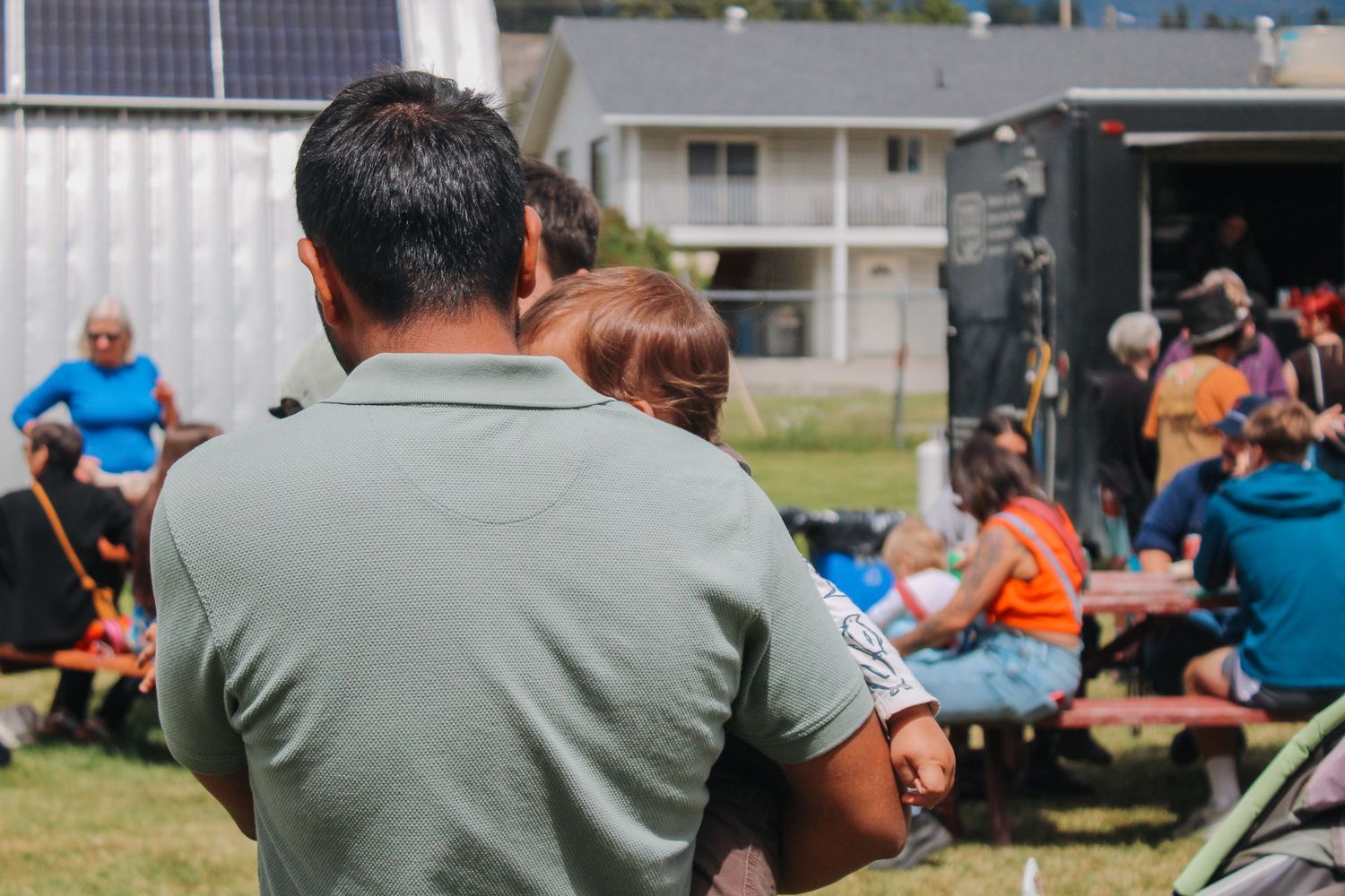 Man holding a child at an outdoor community gathering event on a sunny day.