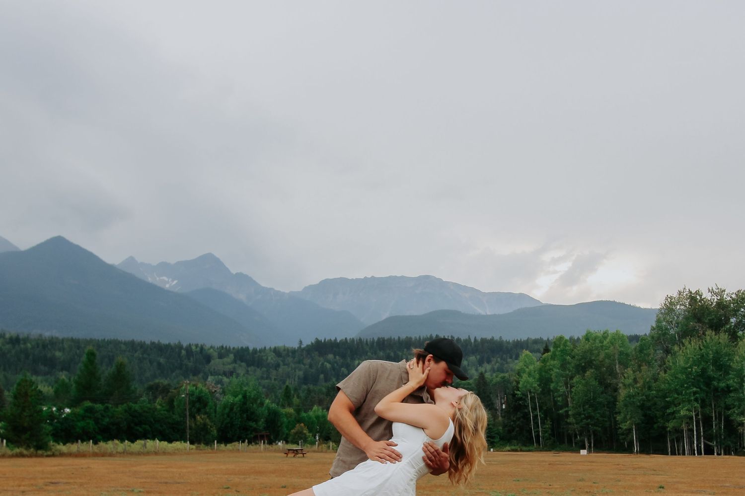 Couple sharing a romantic dip kiss in a golden field with misty mountain and forest backdrop.