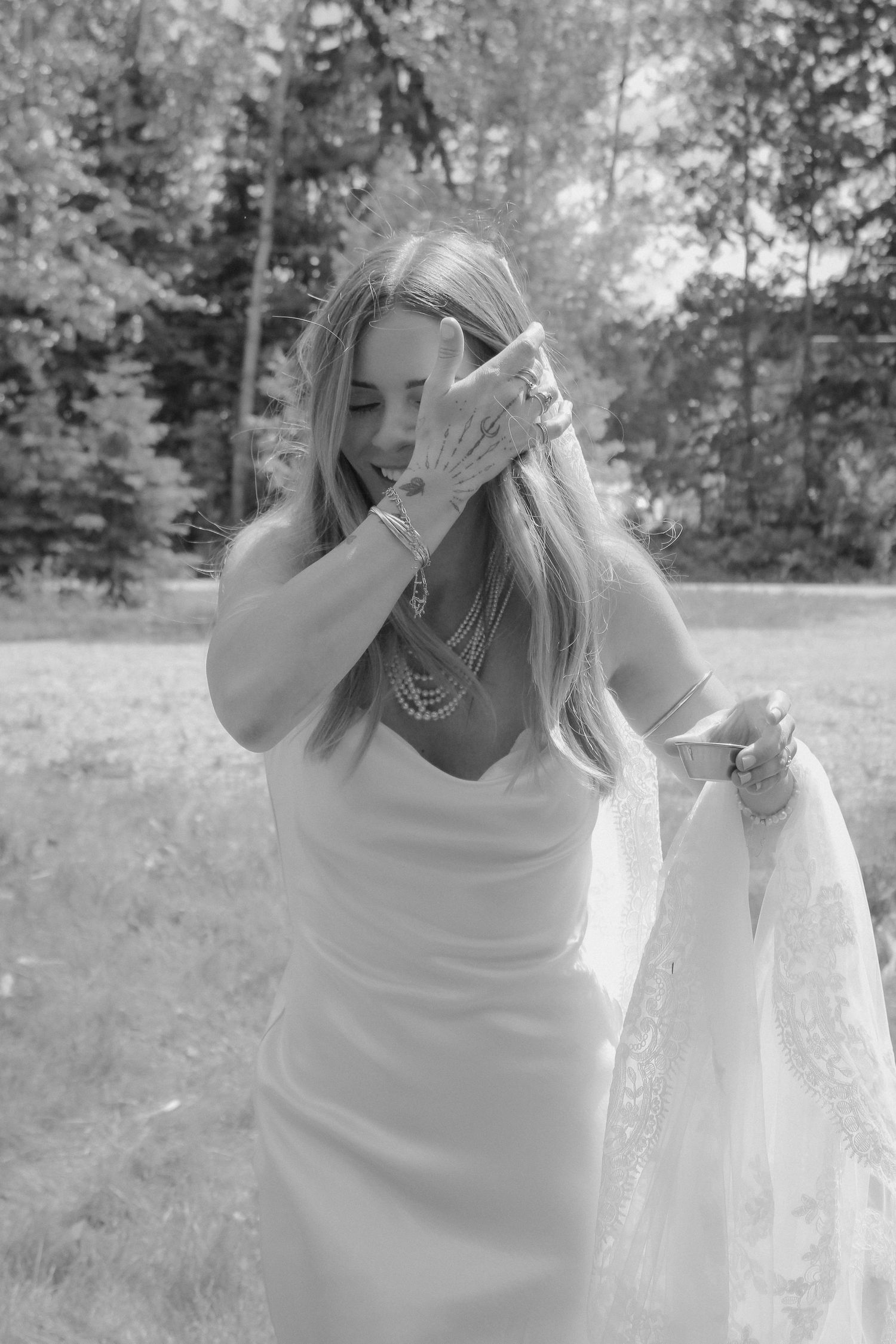 Black and white photo of a bride in a white gown adjusting her hair outdoors, wearing pearl jewelry.