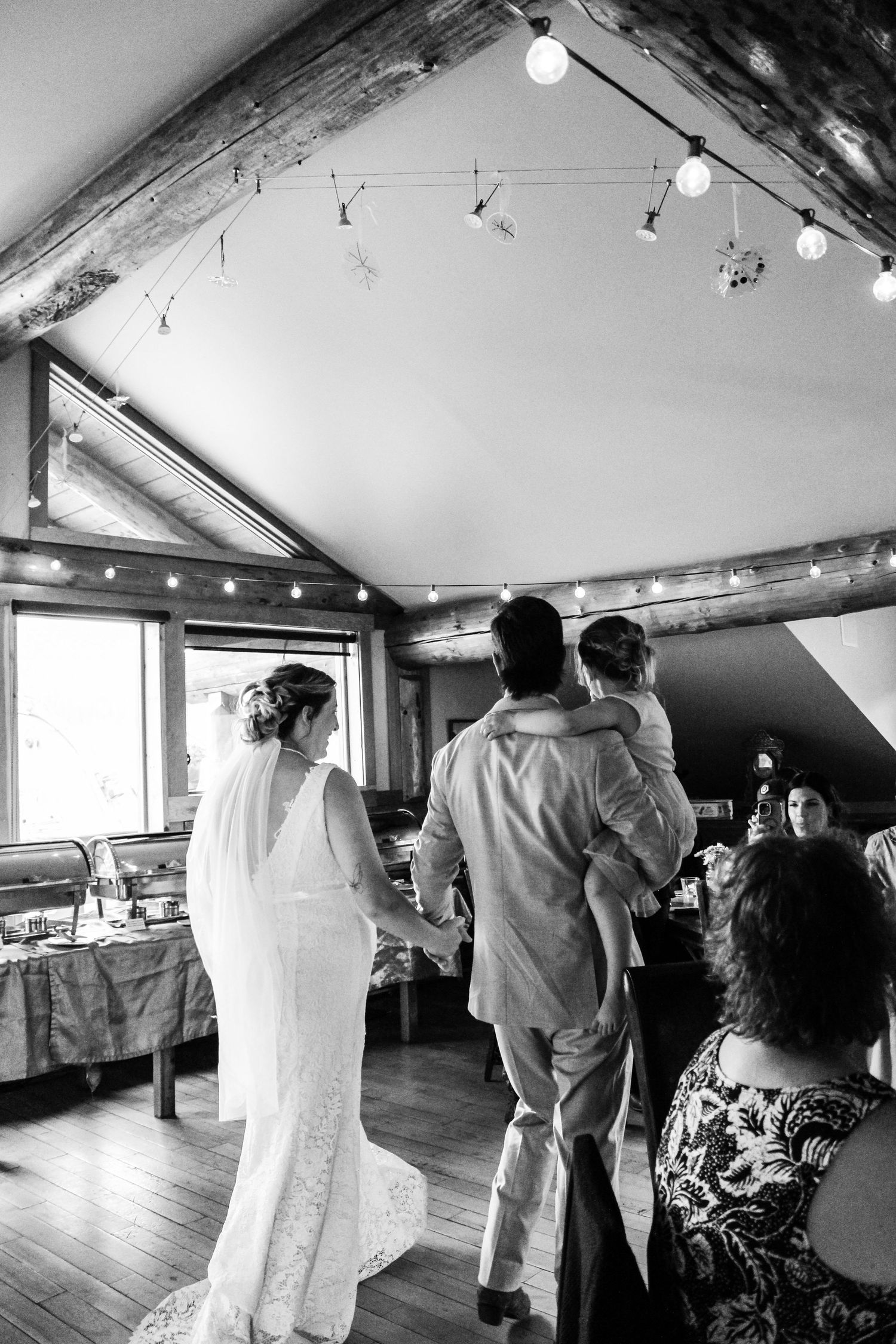 Black and white wedding reception scene showing guests mingling in a rustic indoor space.
