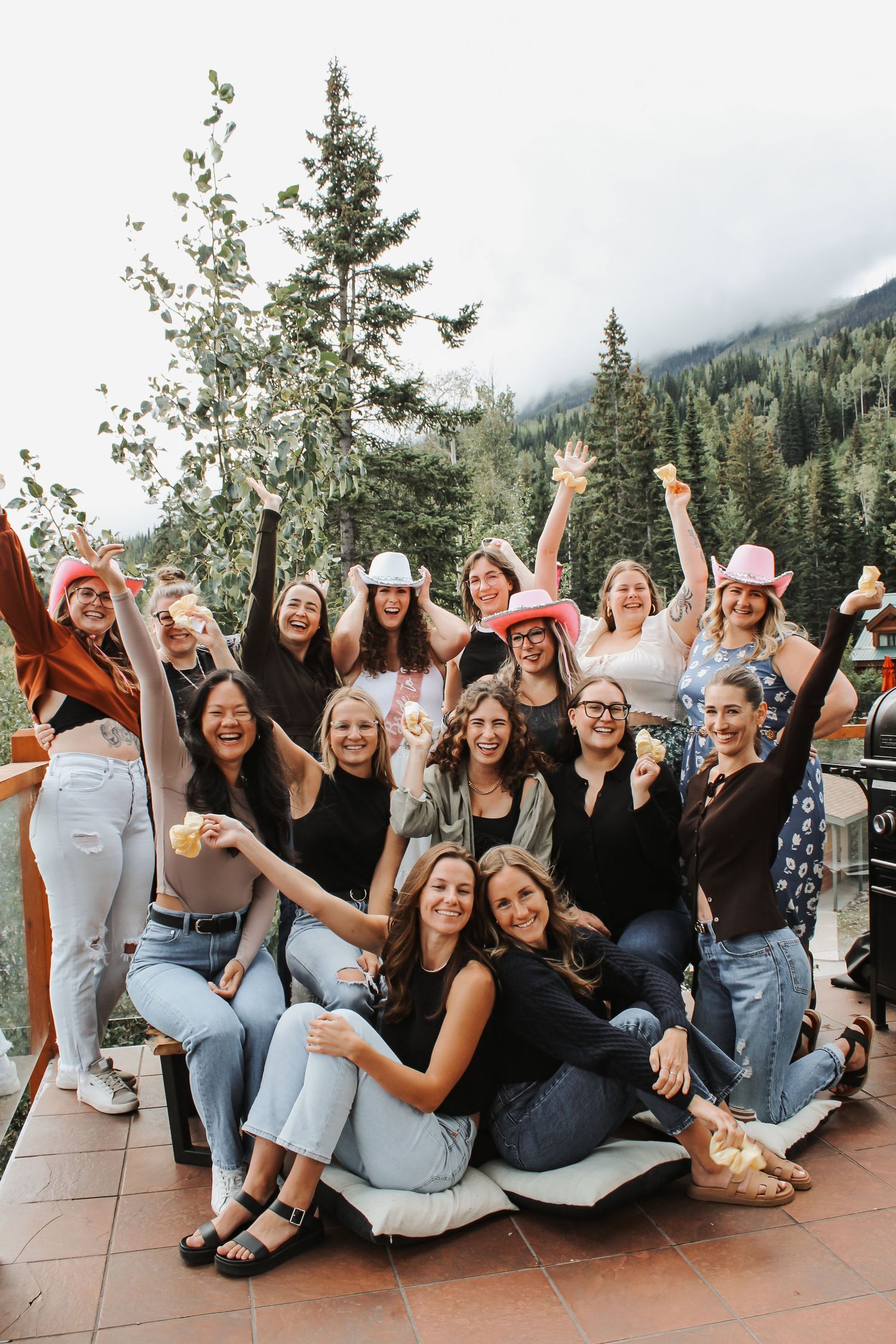 Group of women celebrating a bachelorette party on a mountain resort balcony surrounded by pine trees.