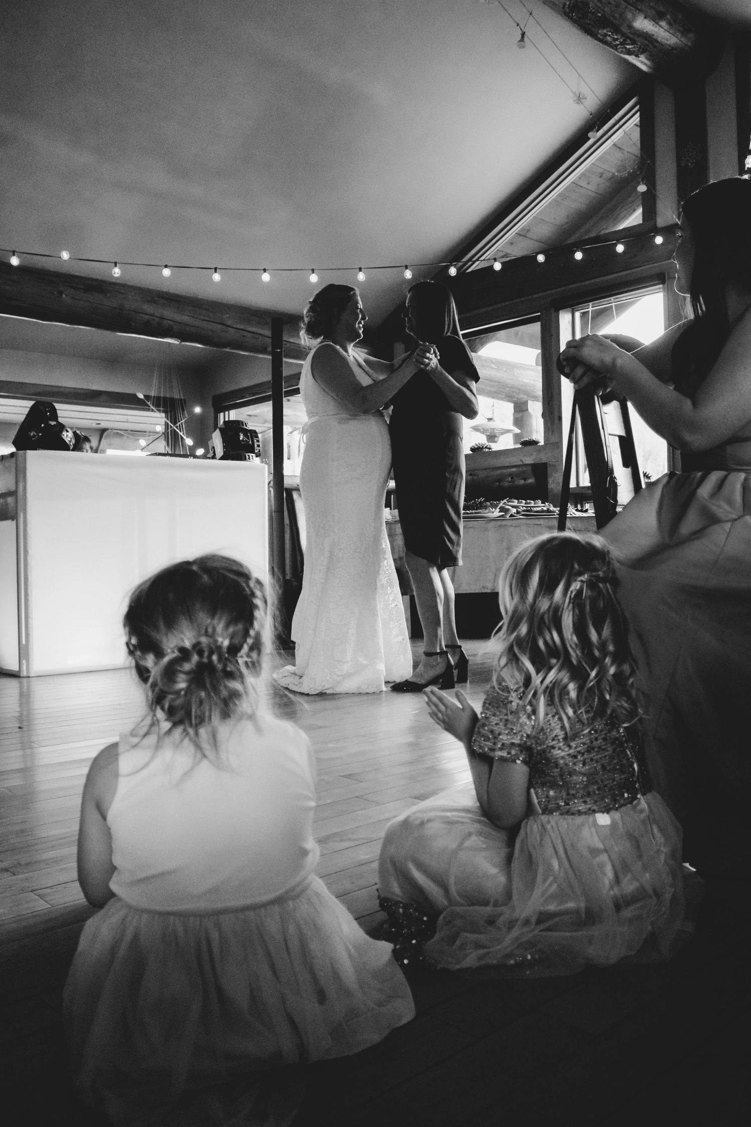 Low angle view of people dancing in white dresses on a wooden floor with string lights overhead.